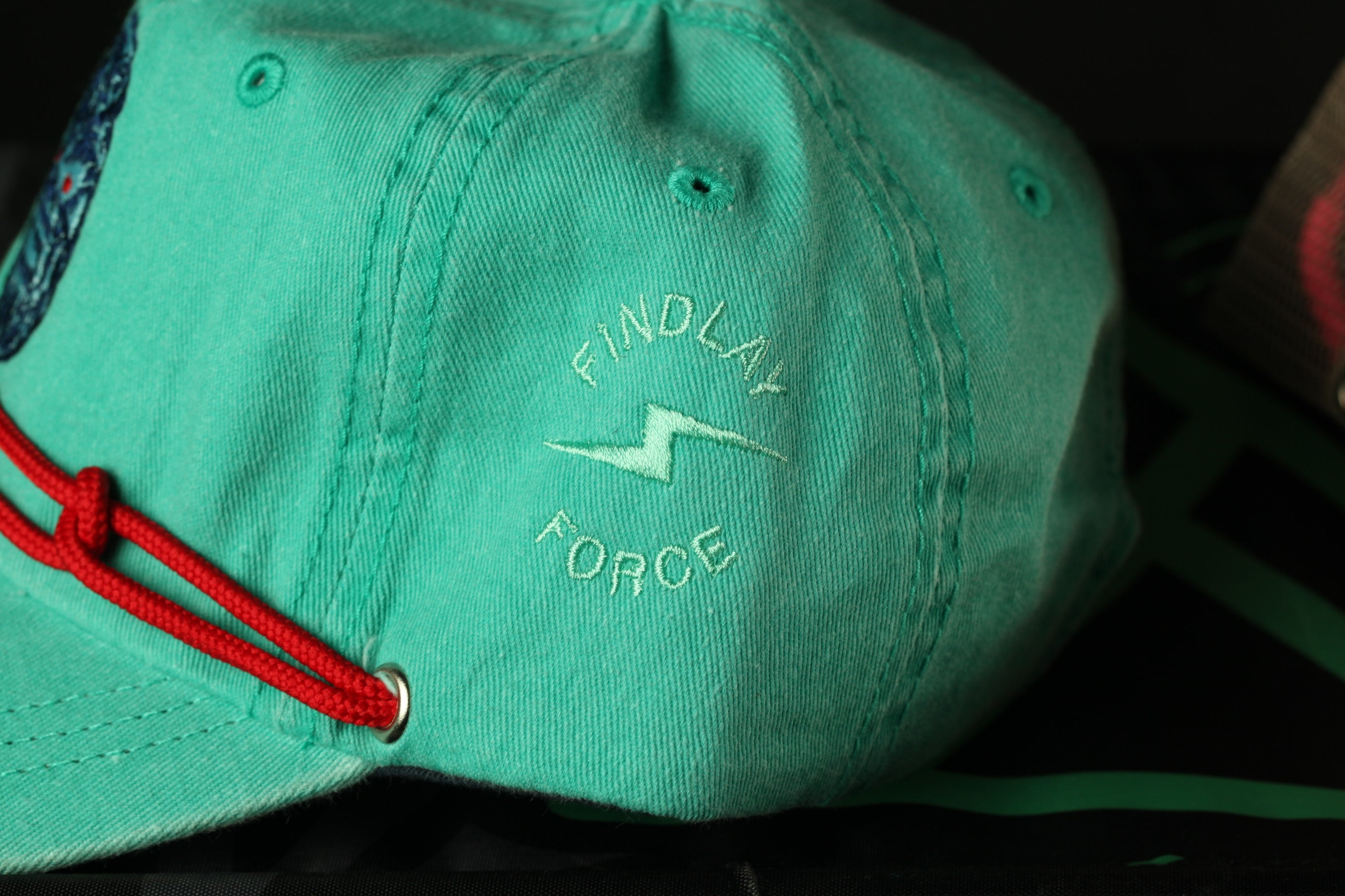 A close-up view of a turquoise baseball cap with a red string and a logo on the front.