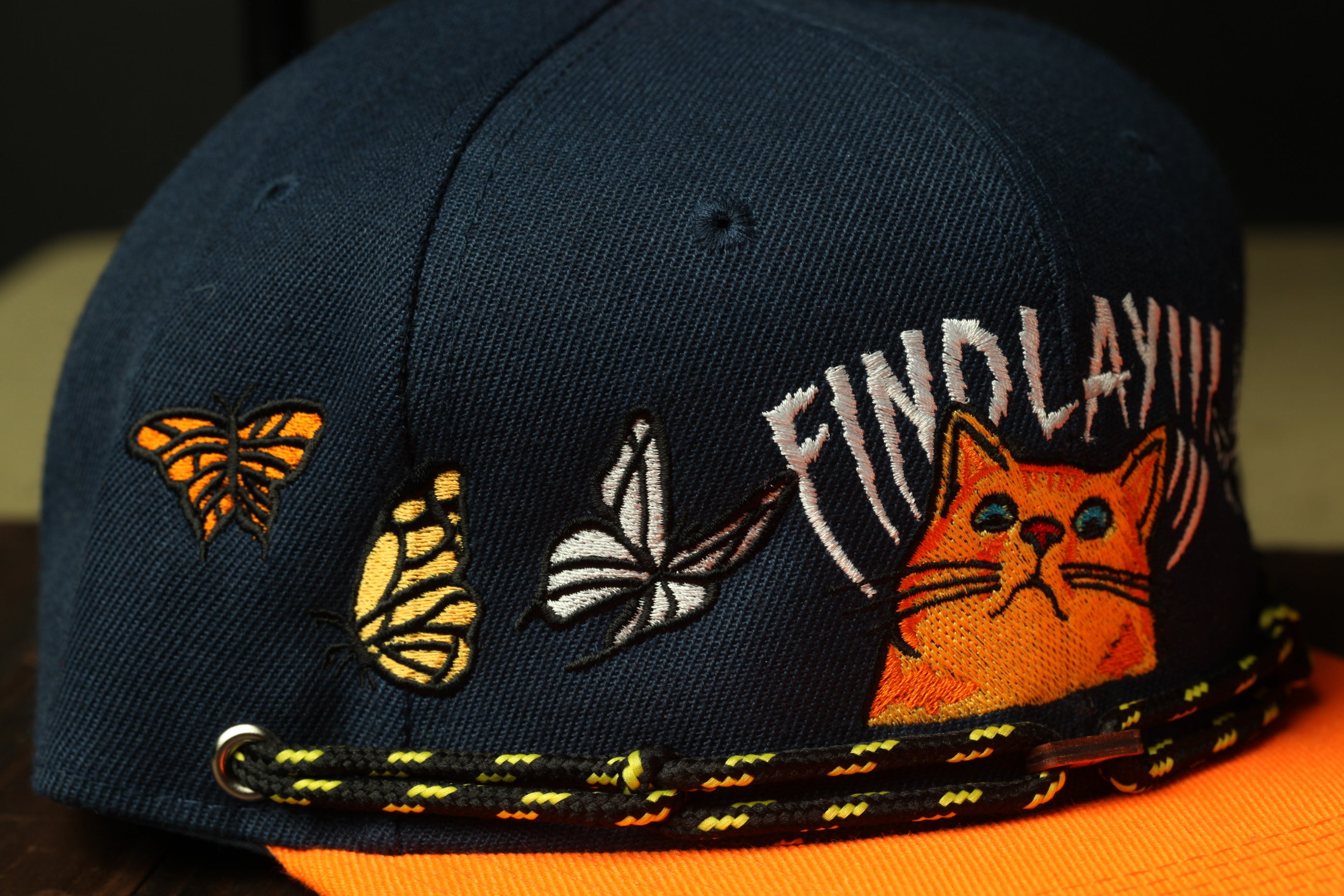 A close-up view of a black baseball cap with various embroidered designs, including a cat, butterflies, and text.