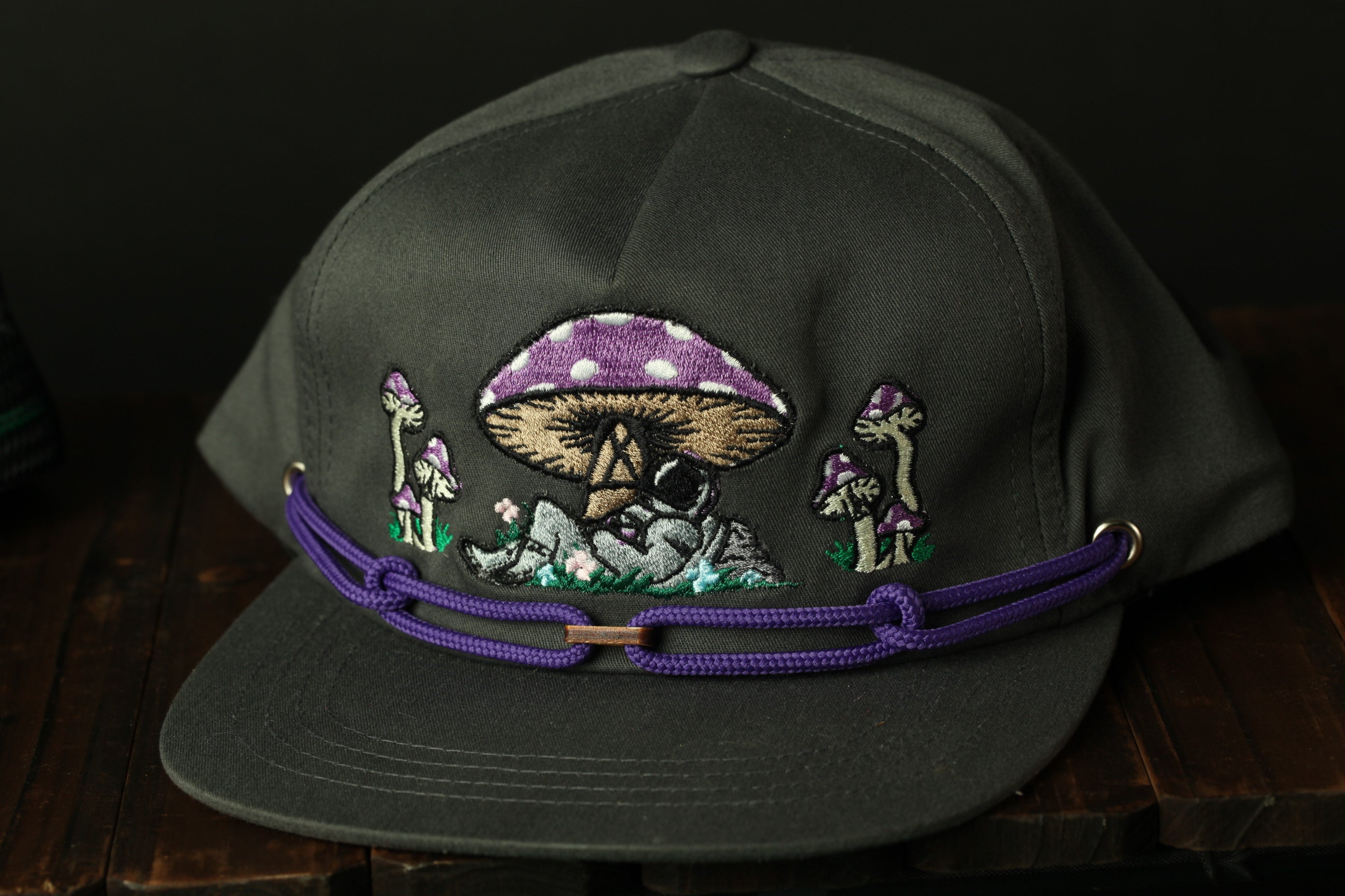A black baseball cap with a purple and white mushroom design and other mushrooms on a wooden surface.
