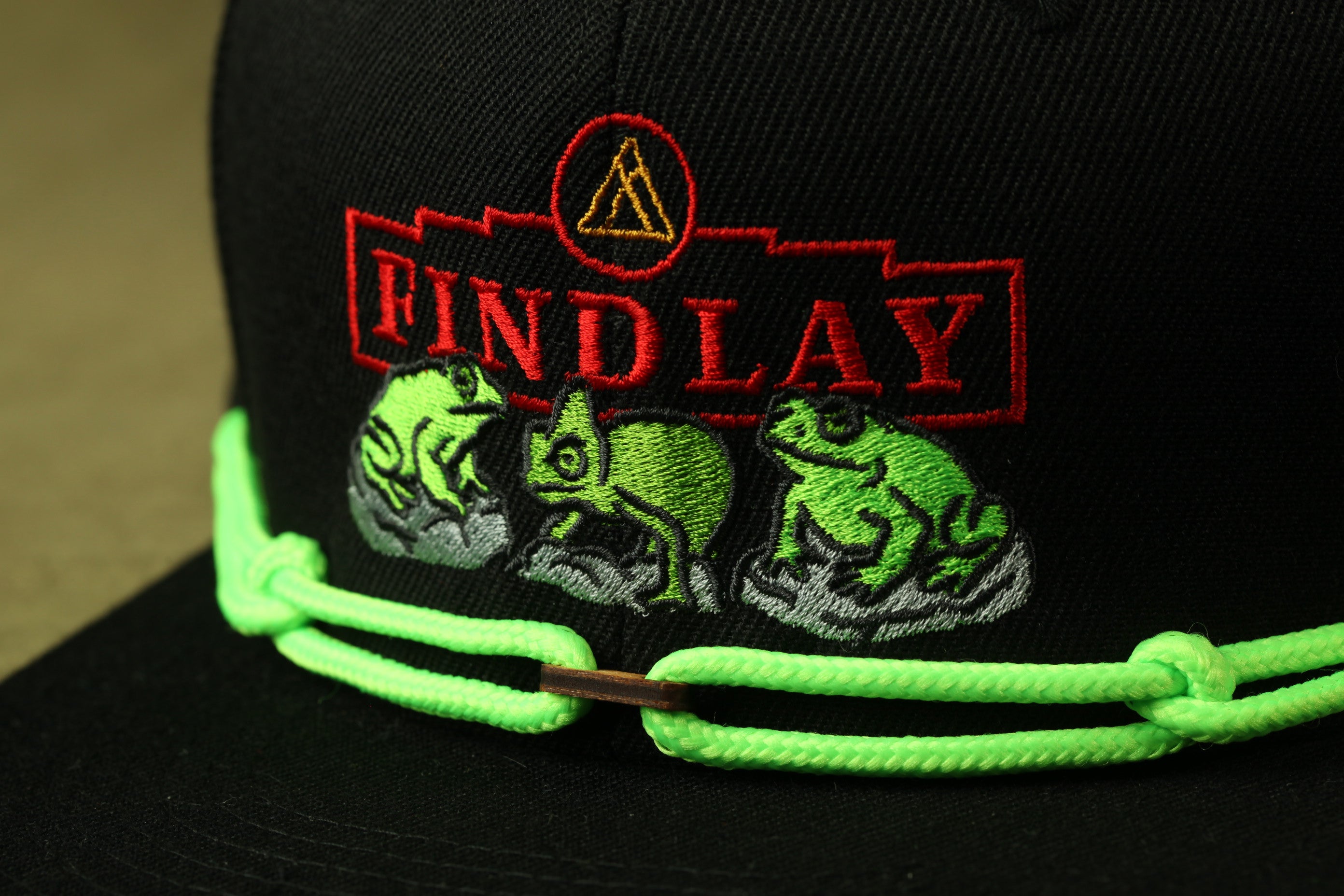 A close-up view of a black baseball cap with a green rope and a logo featuring three green frogs.