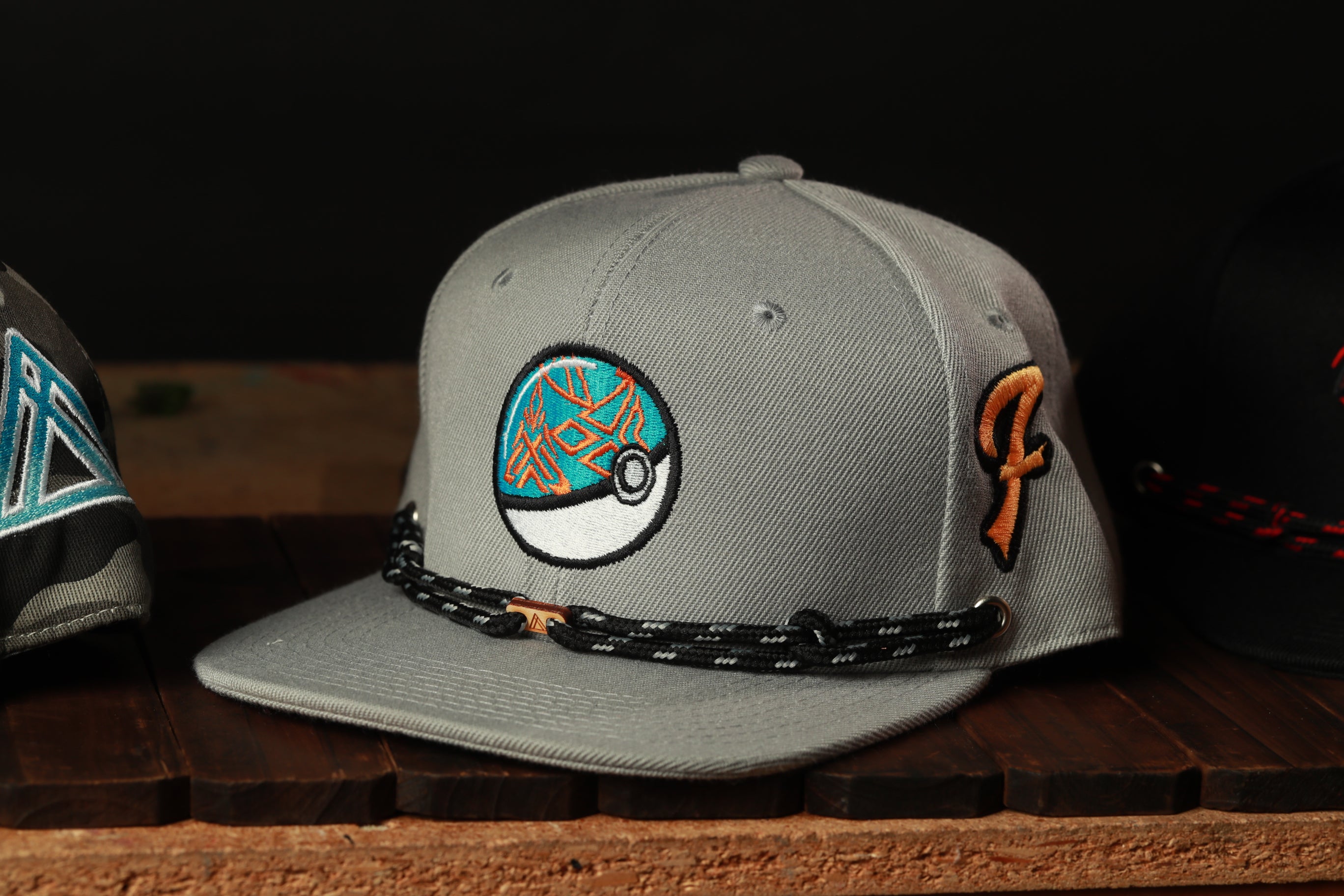 A gray baseball cap with a Pokemon-inspired design, including a blue and orange Pokemon ball, is displayed on a wooden surface.