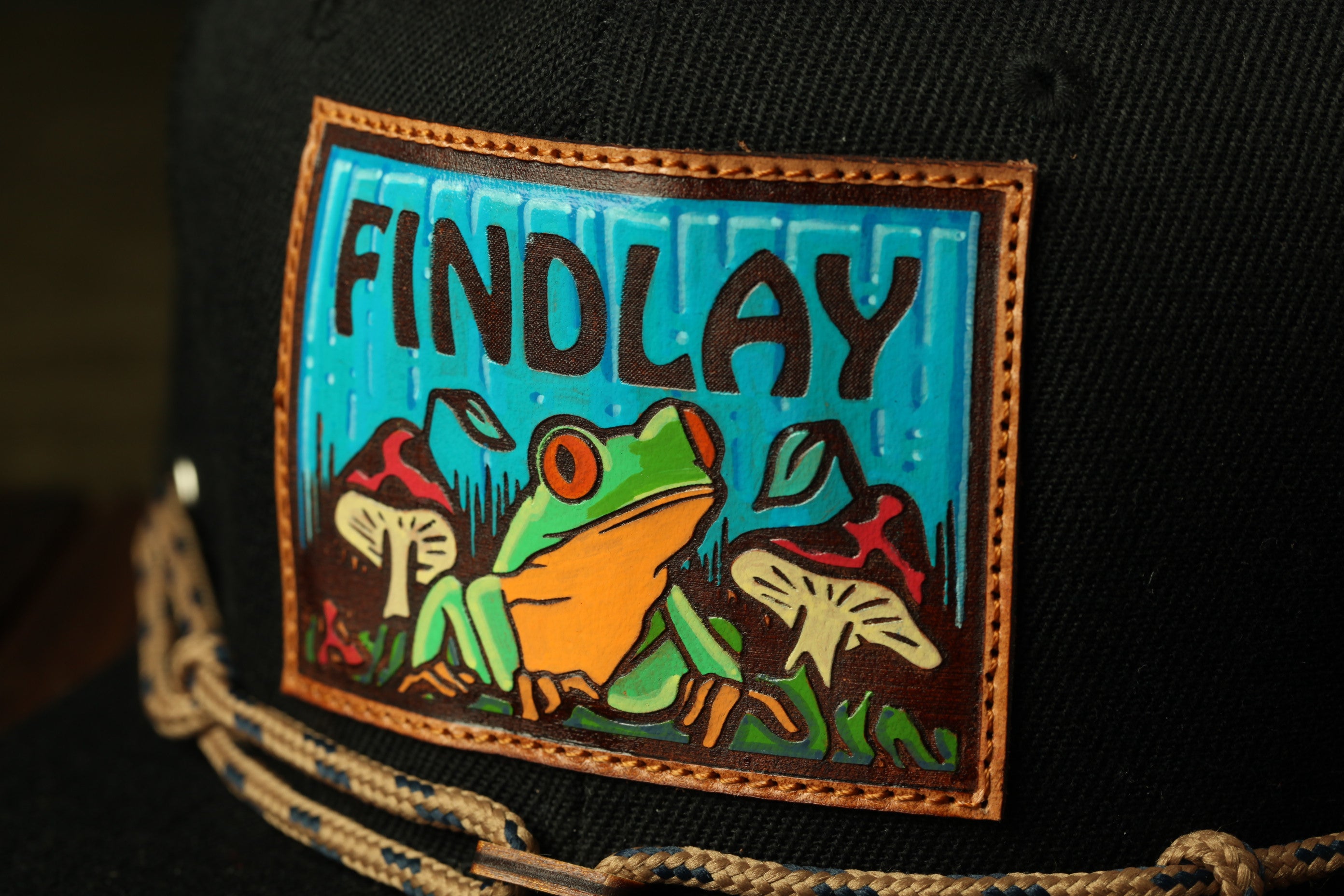 A close-up of a black baseball cap with a colorful patch featuring a frog and mushrooms.