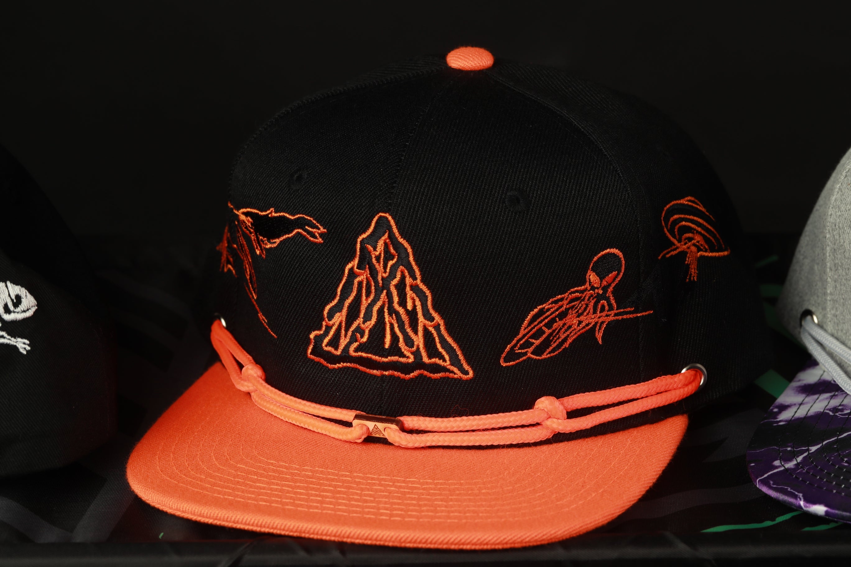 A black baseball cap with orange and orange-like designs on it, displayed on a shelf among other hats.