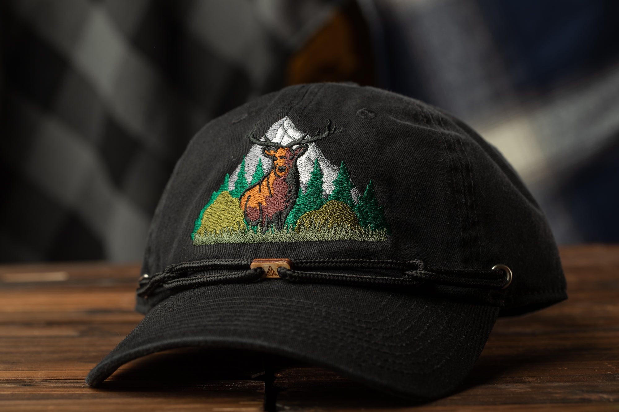 A black baseball cap with an embroidered design of a deer standing in a grassy field with mountains in the background.