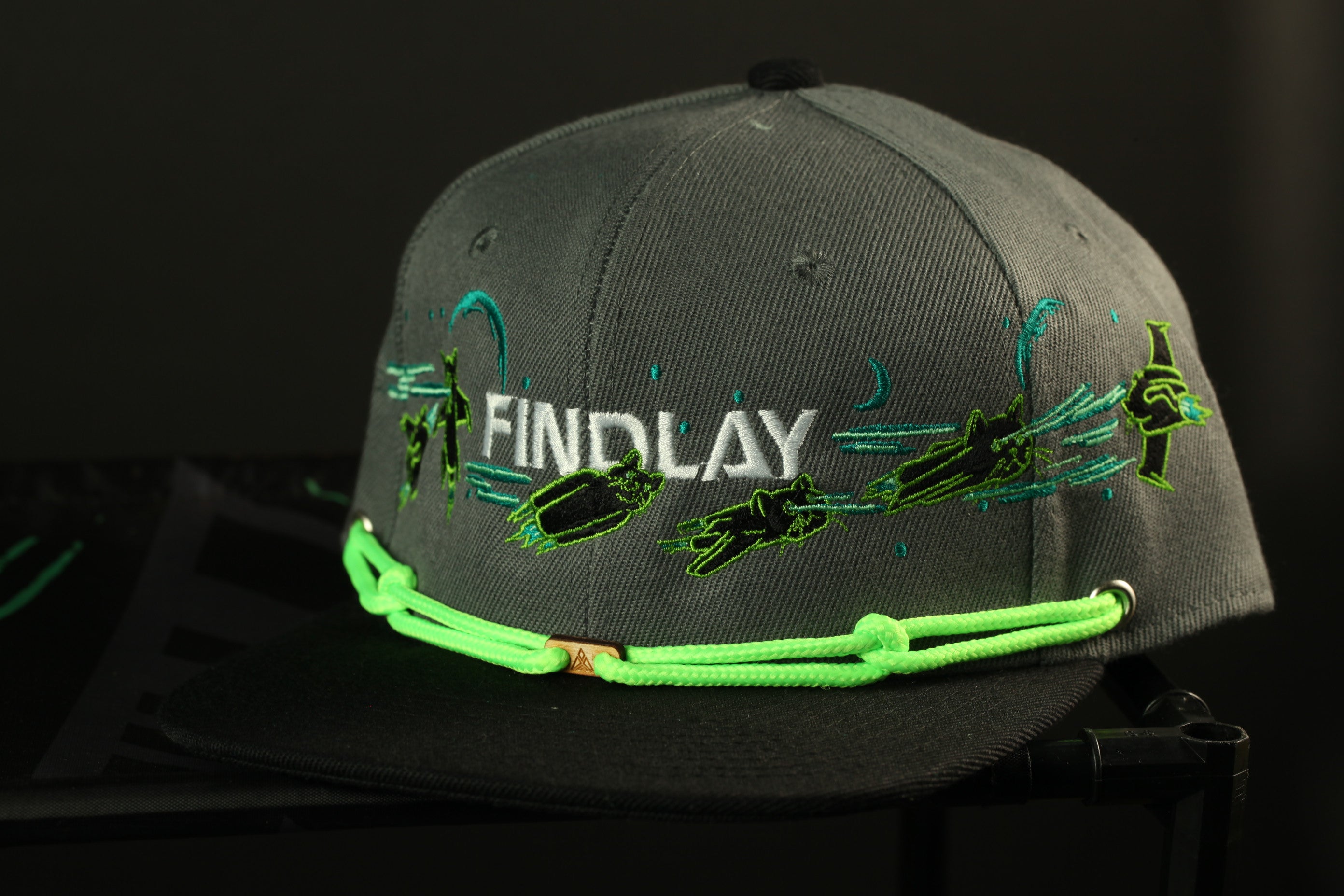 A gray baseball cap with the word "FINDLAY" embroidered on it, along with various green and black designs.