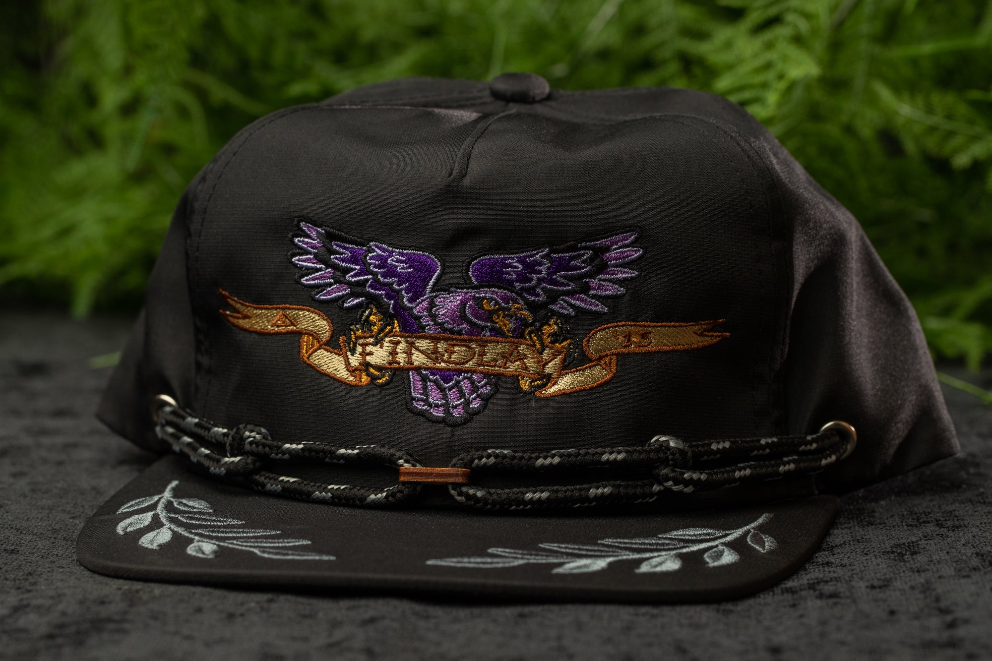 A black baseball cap with a purple eagle design and a banner that says "HINDRIX" on it, placed on a surface with greenery in the background.