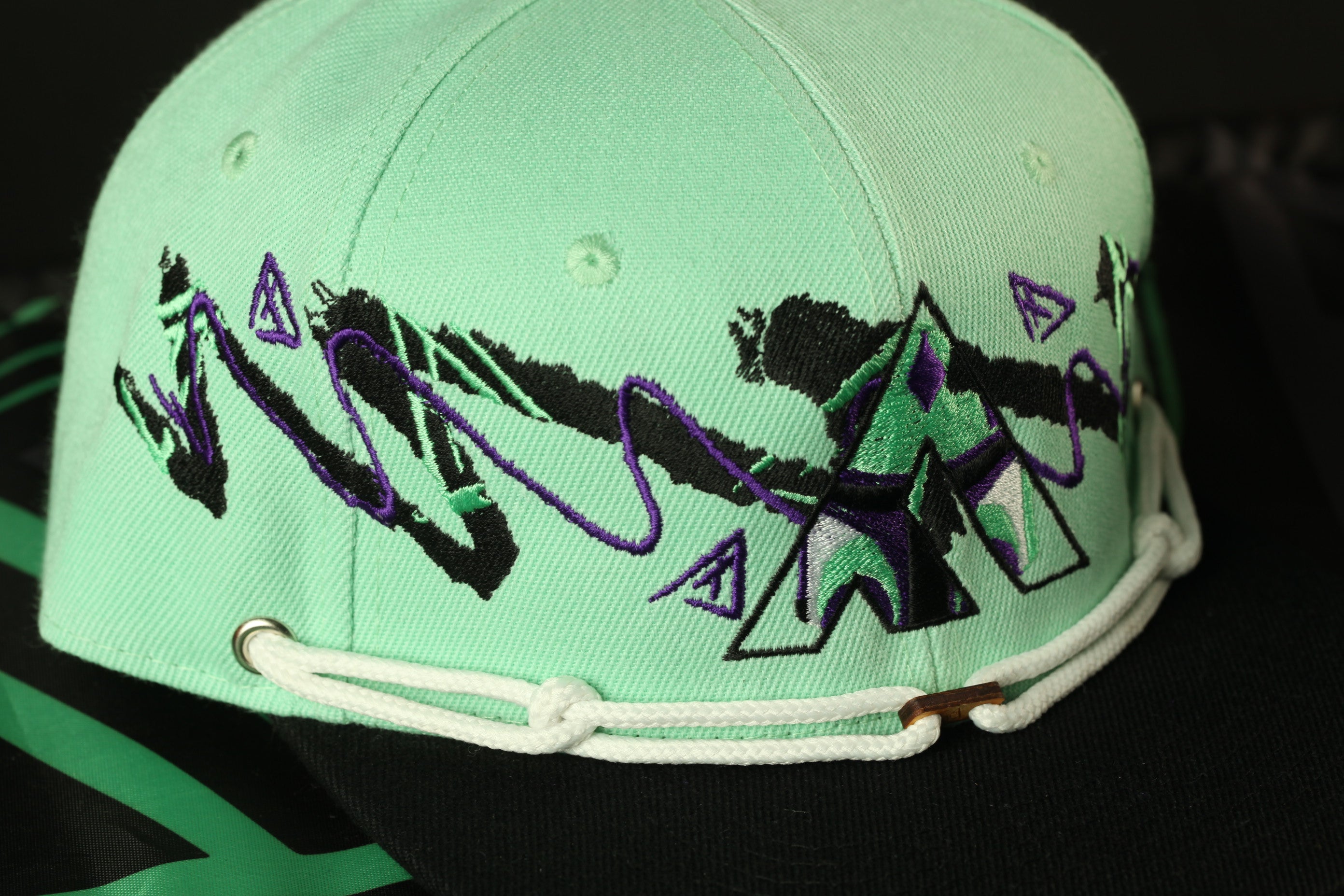 A close-up image of a green baseball cap with a purple and black graphic design on the front.