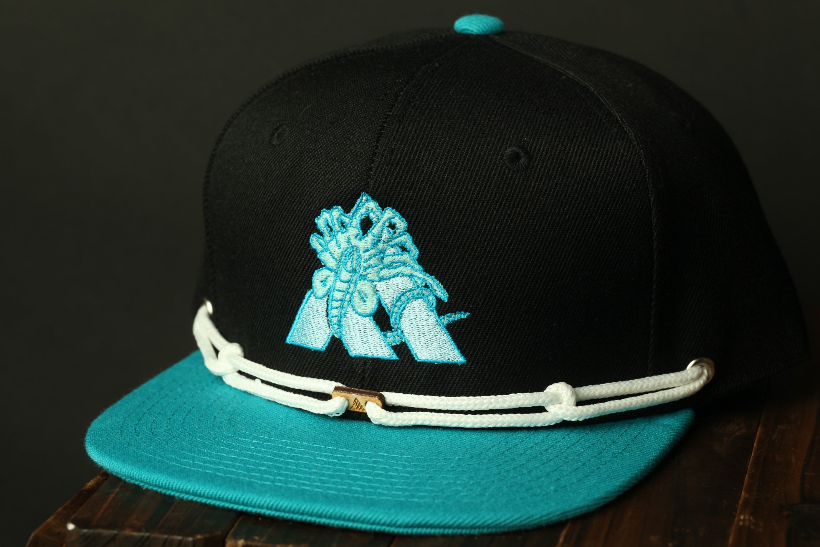 A black baseball cap with a teal brim and white lace trim, featuring a blue logo on the front.