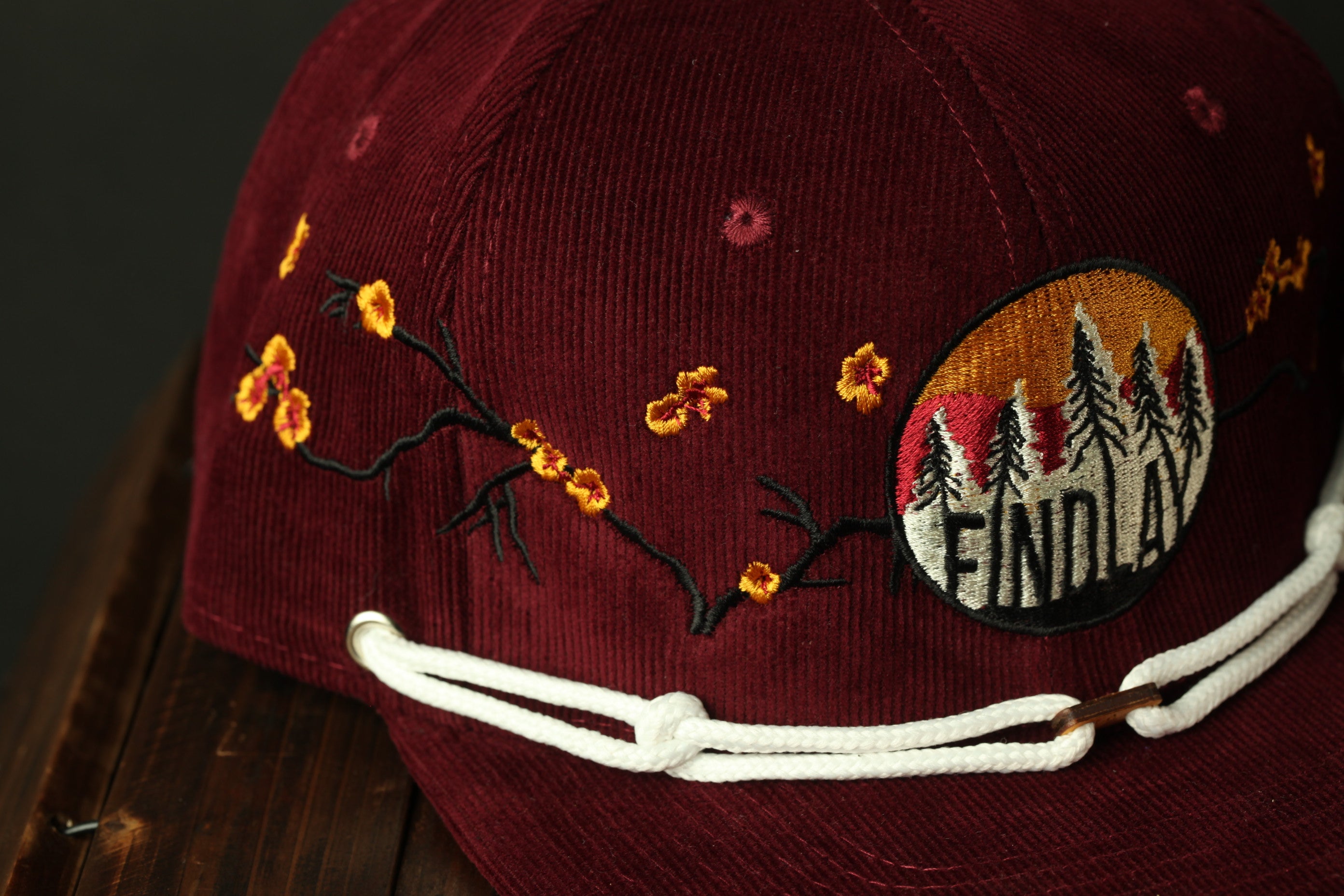 A burgundy baseball cap with a floral embroidered design and a patch on the front that says "FIND LAY".