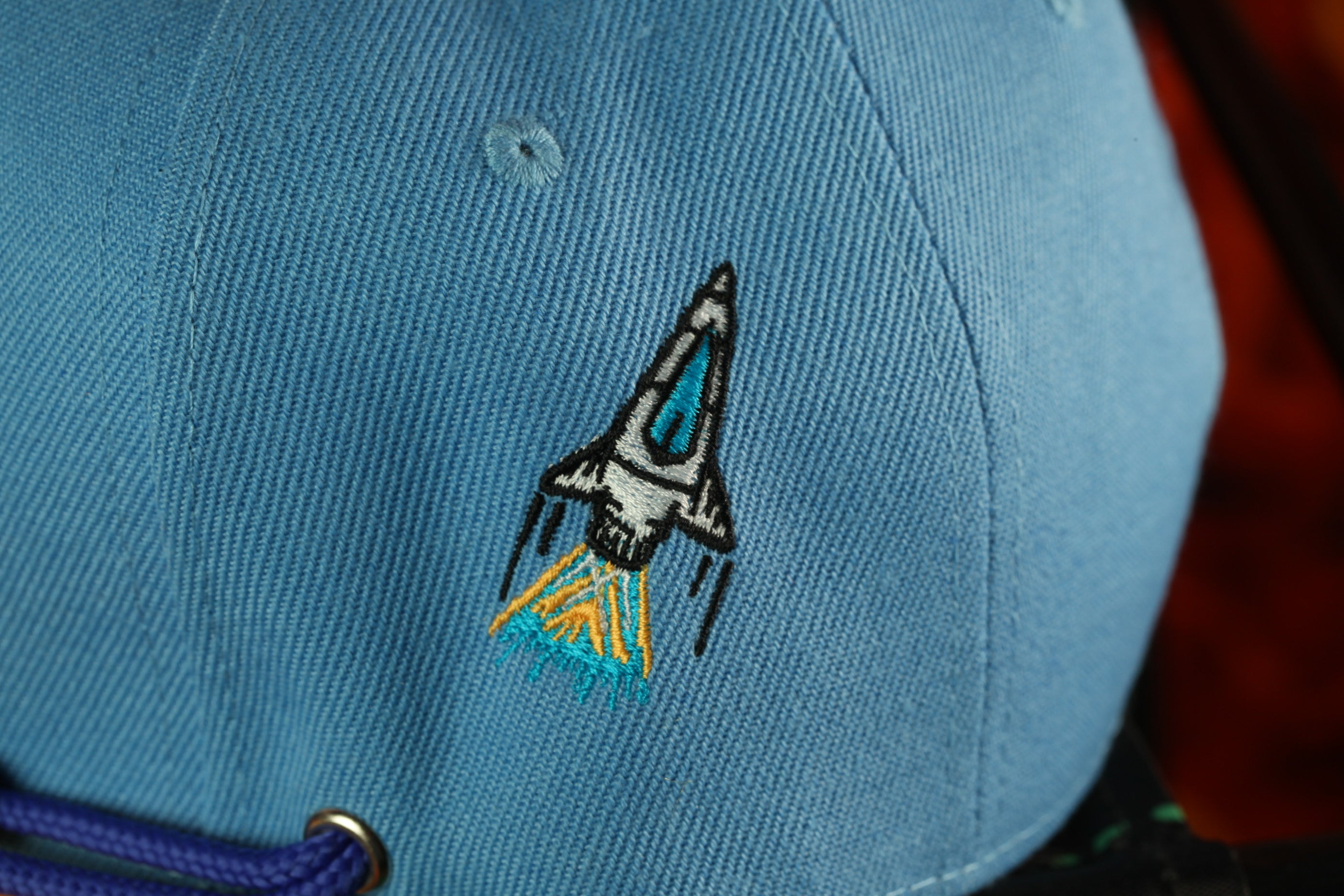 A close-up view of a blue baseball cap with a small embroidered rocket ship design on the front.