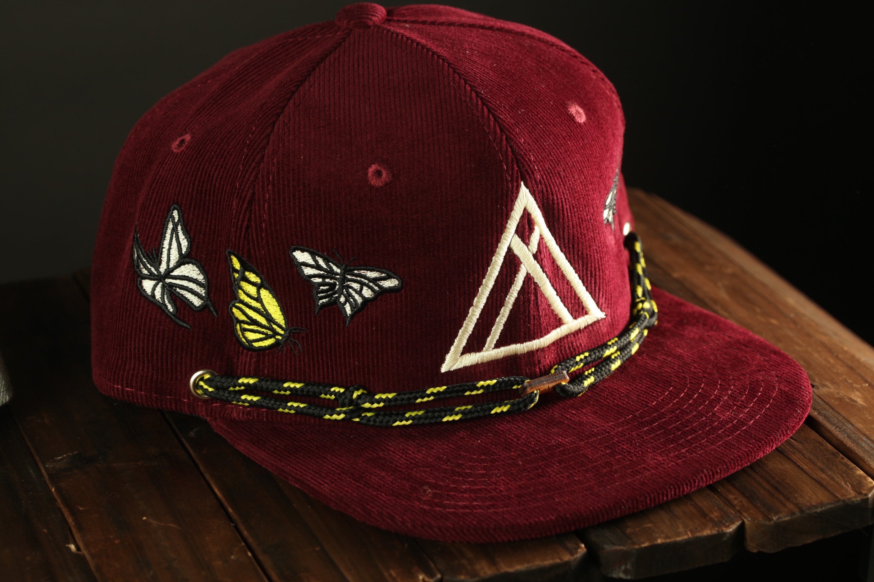 A burgundy baseball cap with a white triangle design and butterfly embroidery, resting on a wooden surface.