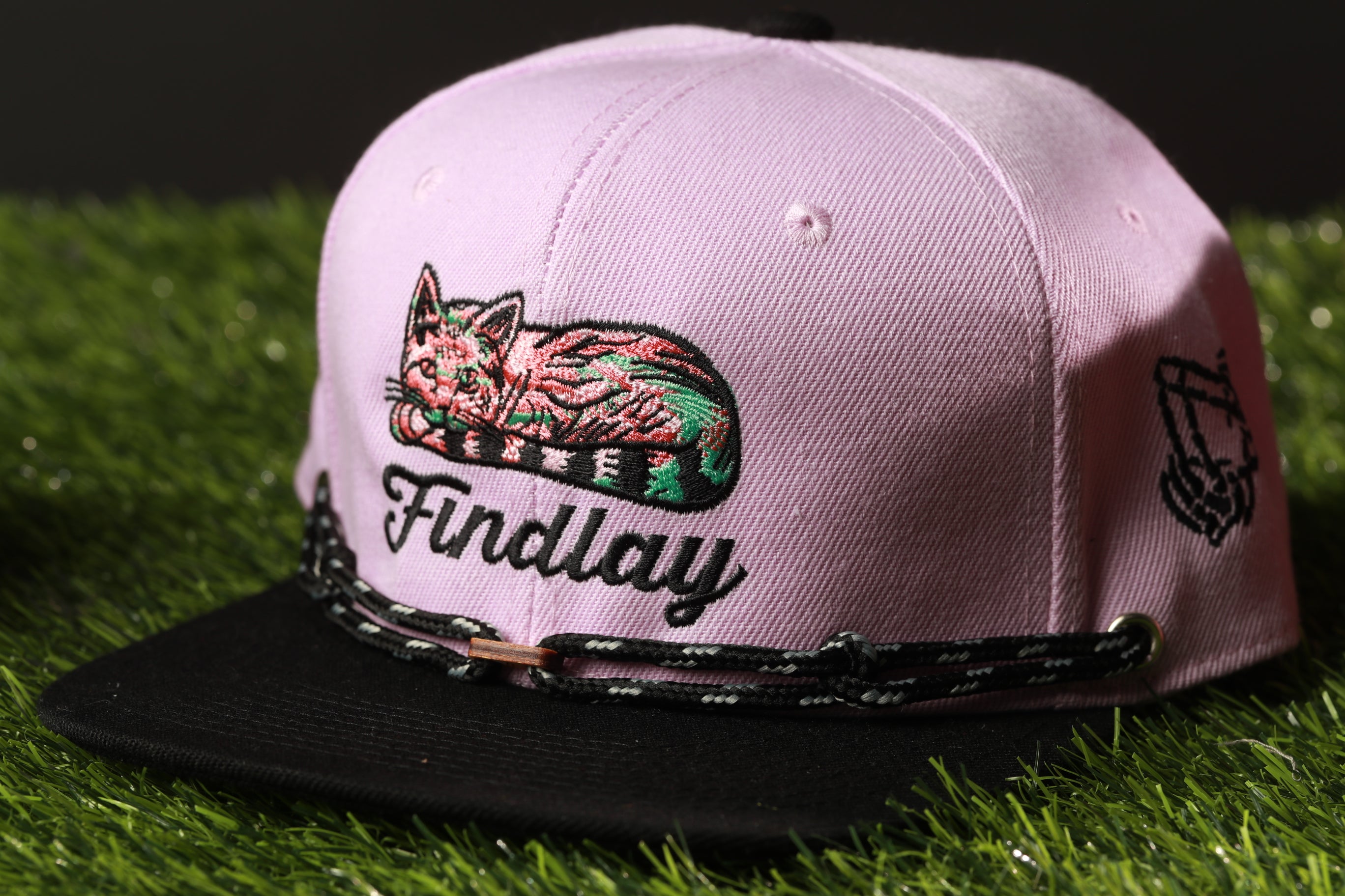 A pink baseball cap with a cat embroidered on it, sitting on a grassy surface.