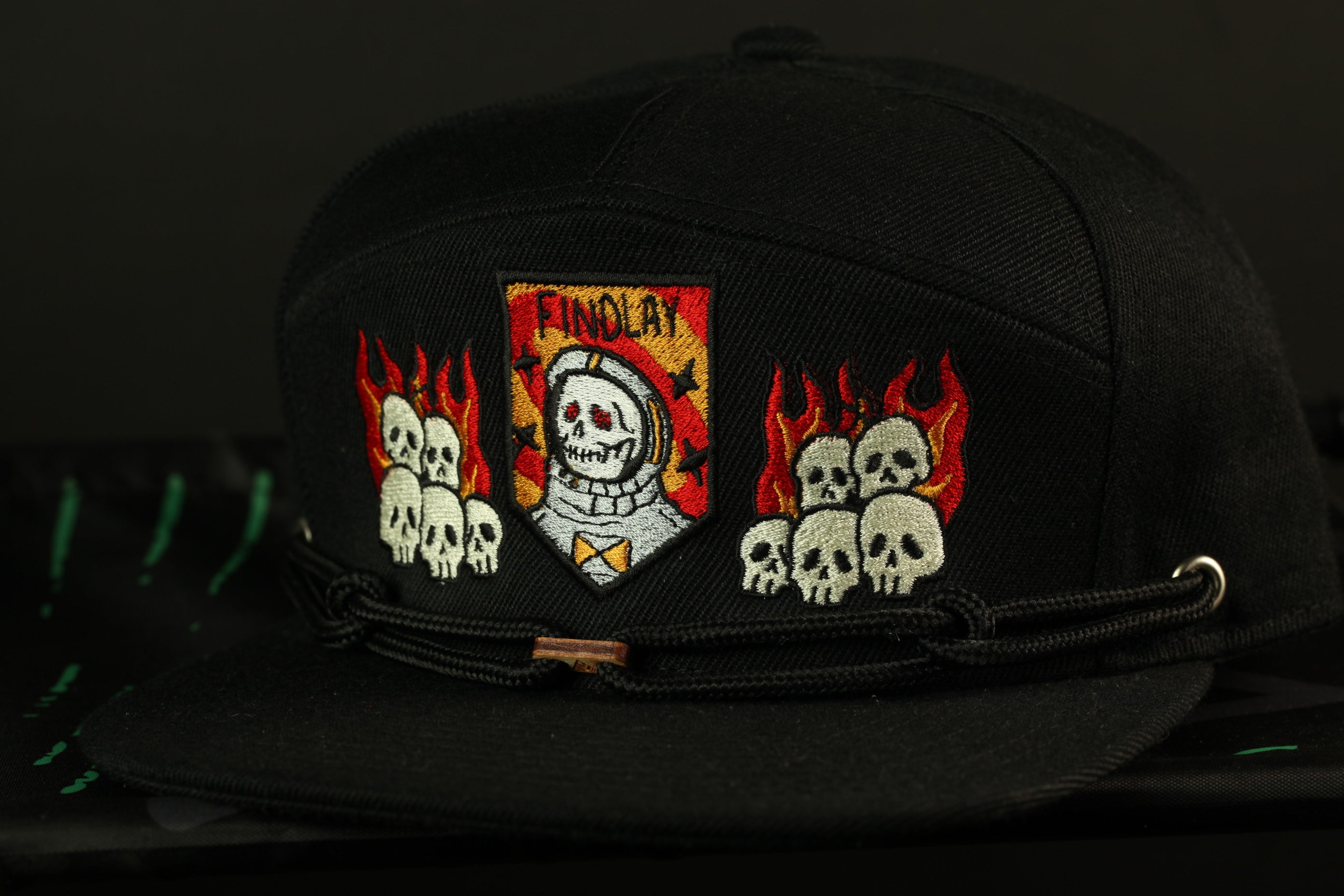 A black baseball cap with a unique design featuring a skull and crossbones motif, along with flames and a shield-shaped emblem.
