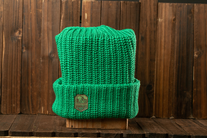 A bright green knitted beanie with a metal patch on the front, placed on a wooden surface against a wooden background.