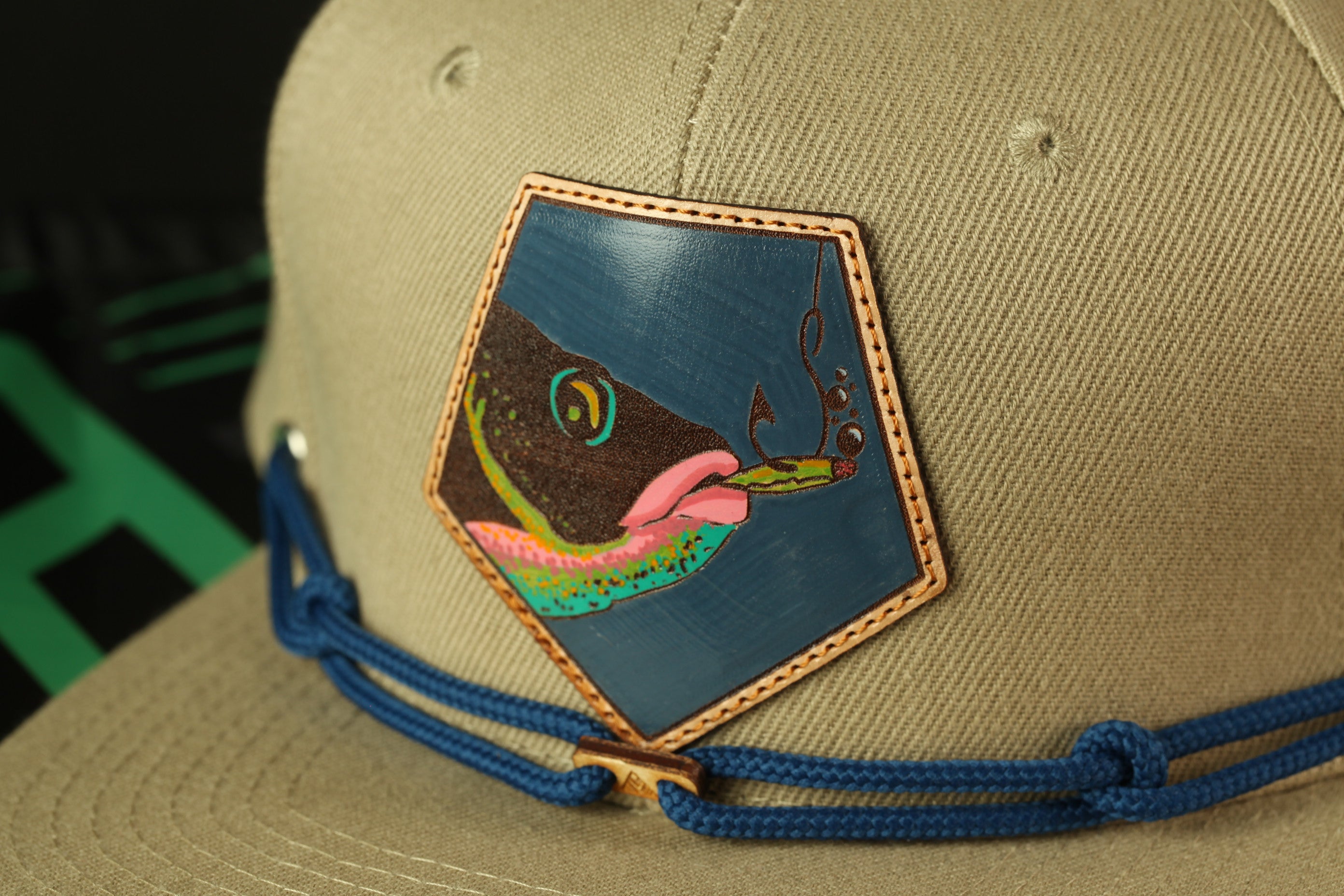 A close-up view of a beige baseball cap with a blue and green fish embroidered on the front.