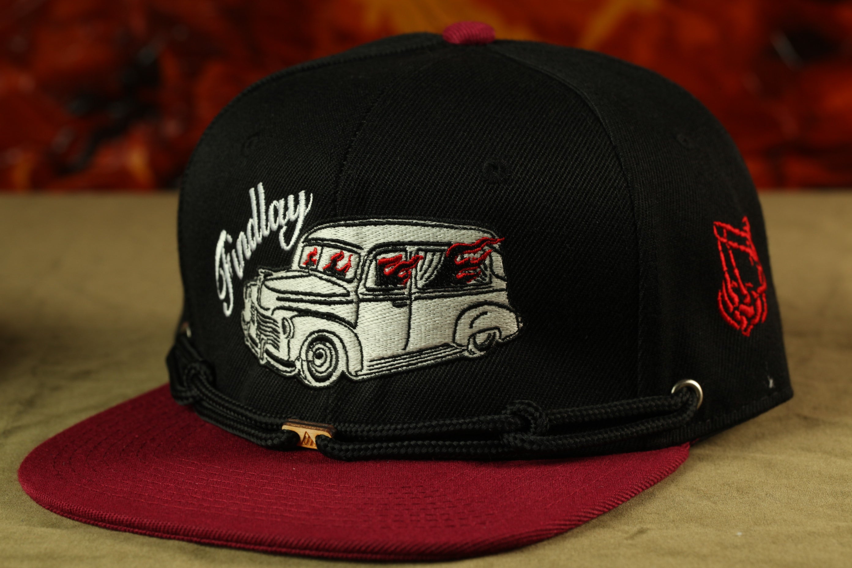 A black baseball cap with a red brim, featuring a white and black illustration of a classic car on the front.