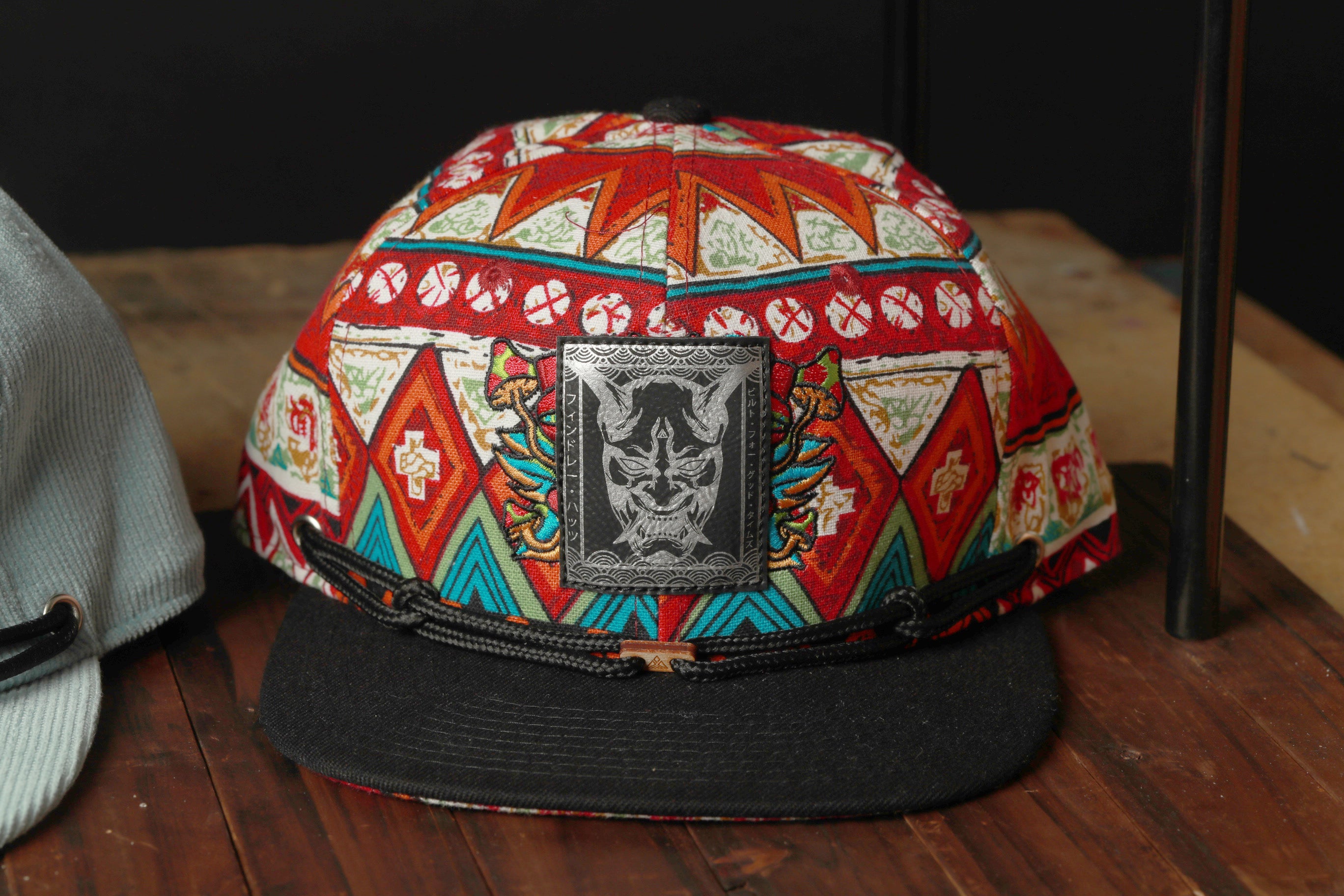 A colorful, patterned baseball cap with a black brim and a metal patch featuring a graphic design is displayed on a wooden surface.