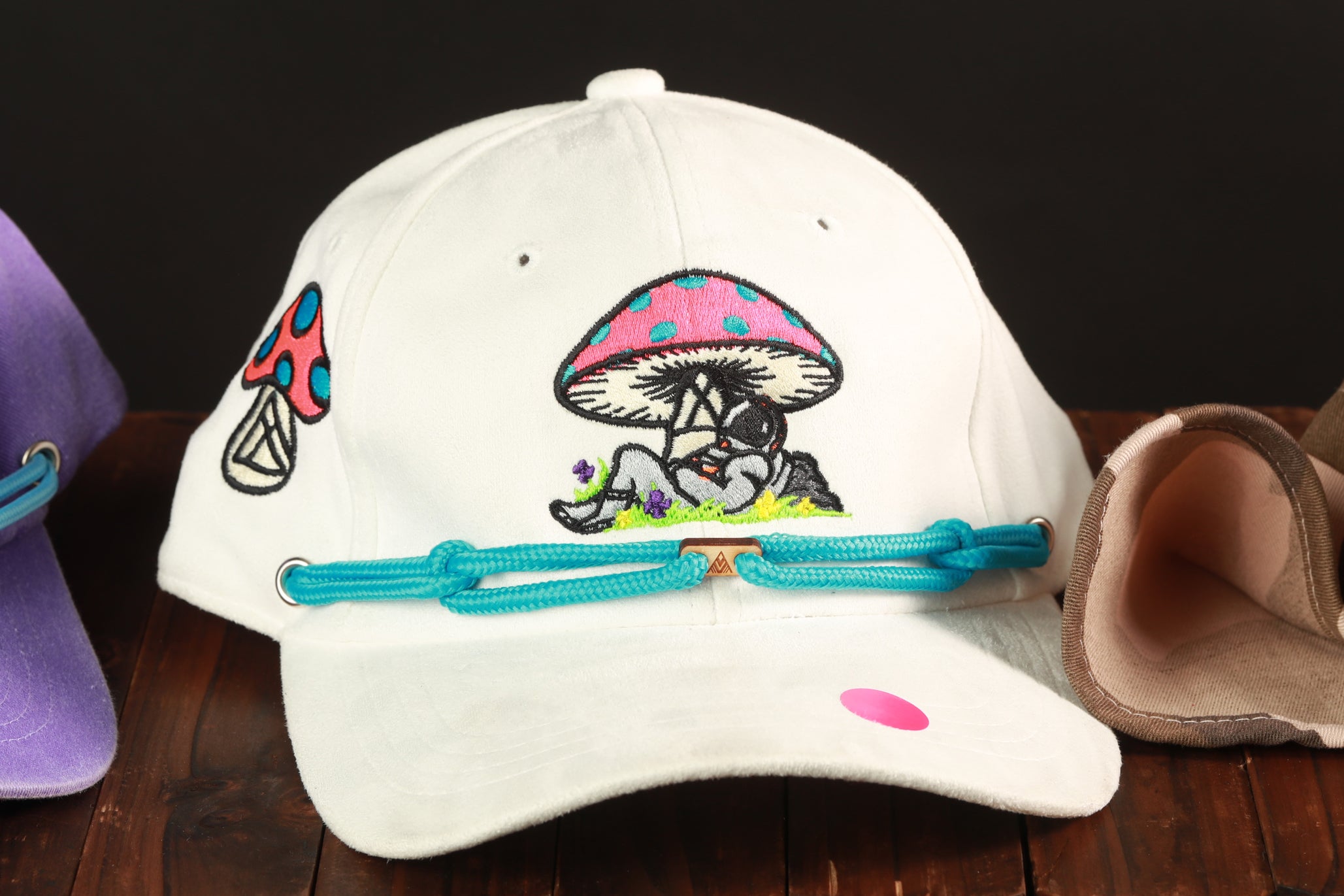 A white baseball cap with a mushroom design and a pink dot on the front, sitting on a wooden surface.