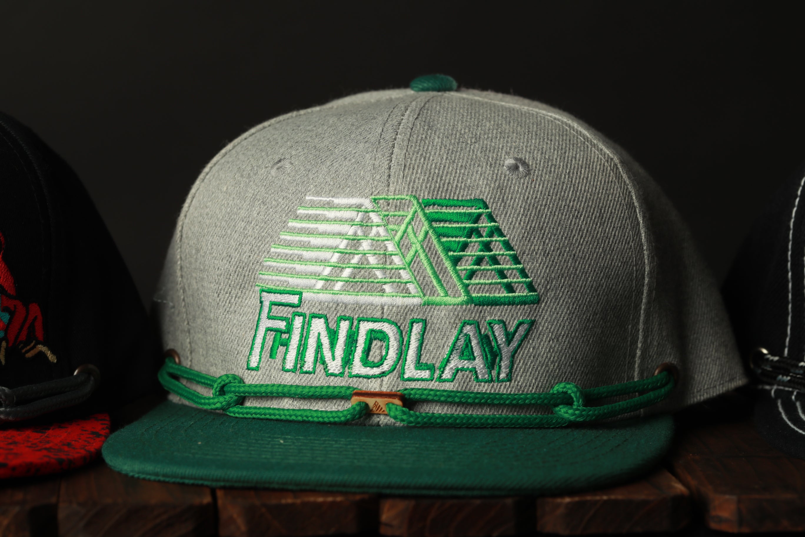 A gray baseball cap with a green band and the word "FINDlay" printed on it, sitting on a wooden surface.