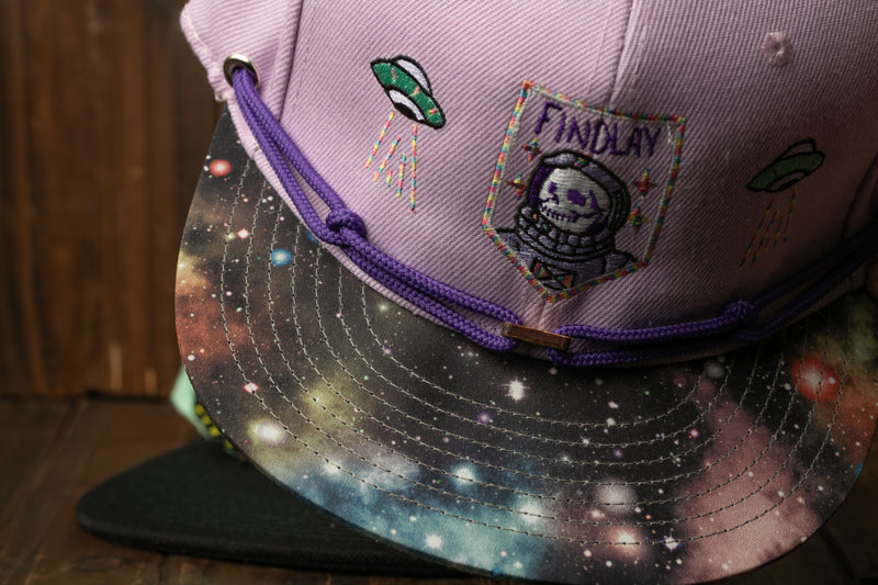 A purple baseball cap with a space-themed design, including a graphic of a skull and a planet, and a purple string.