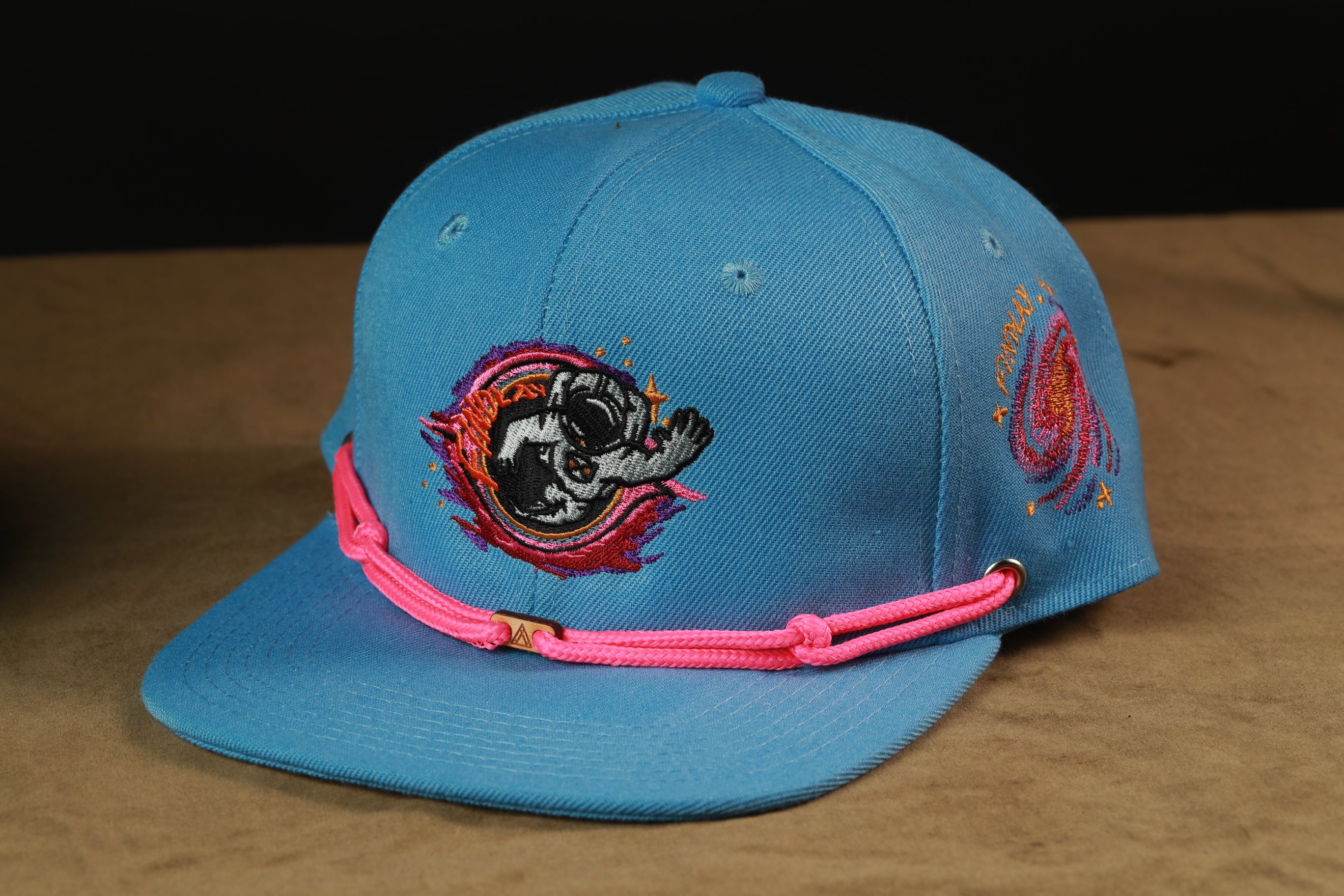 A blue baseball cap with a pink string and a black and white dog embroidered on it, sitting on a brown surface.