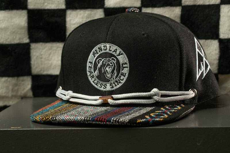 A black baseball cap with a colorful brim and a white logo on the front, sitting on a black surface against a checkered background.