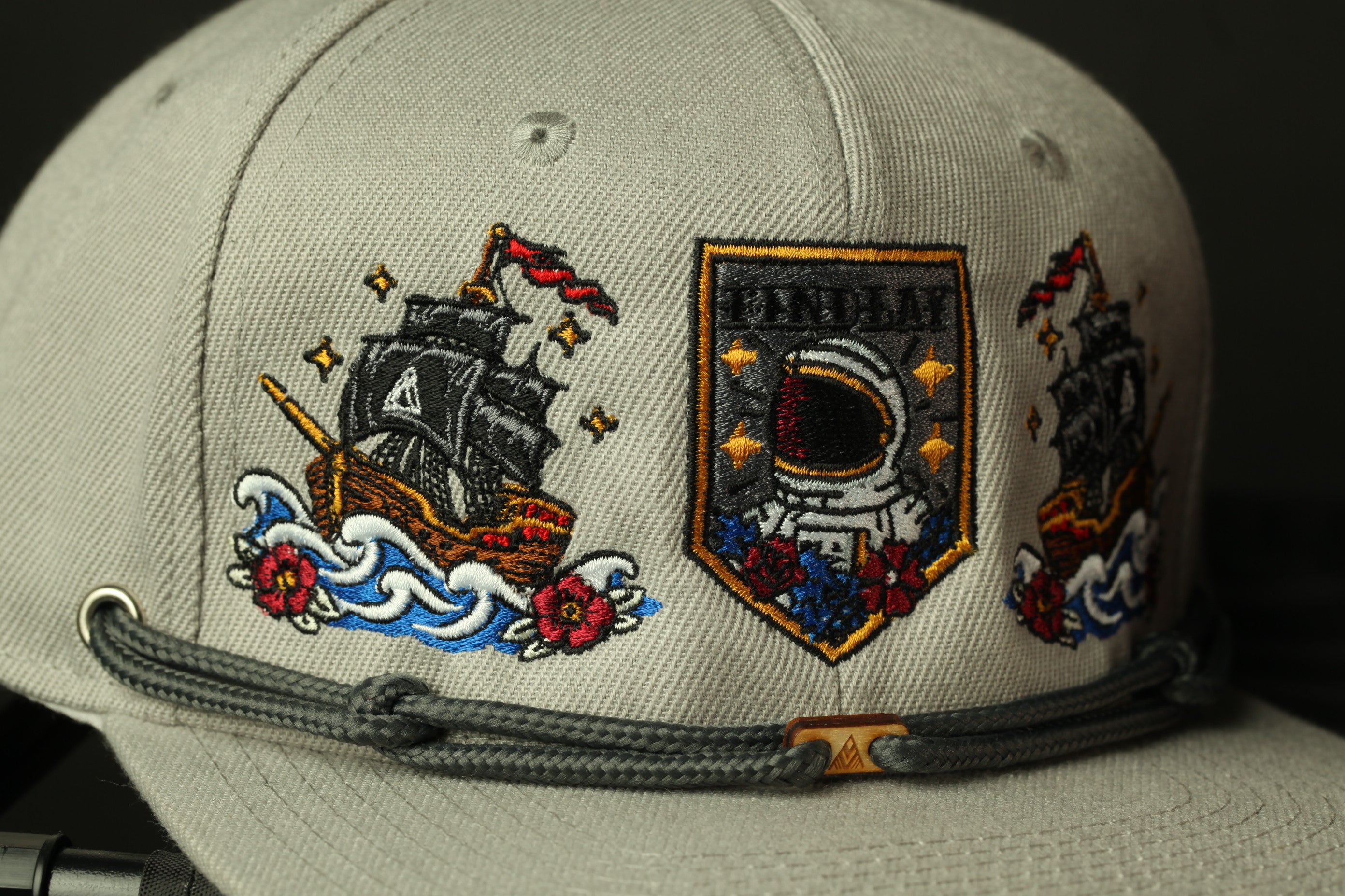 A close-up view of a gray baseball cap with various embroidered designs, including a ship, a person in a spacesuit, and other decorative elements.