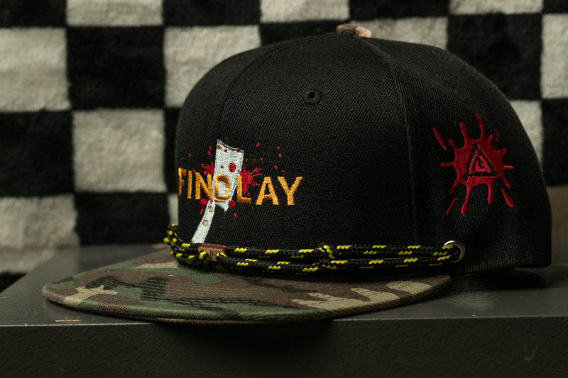 A black baseball cap with a camouflage pattern and a red splatter design on the side, sitting on a surface.