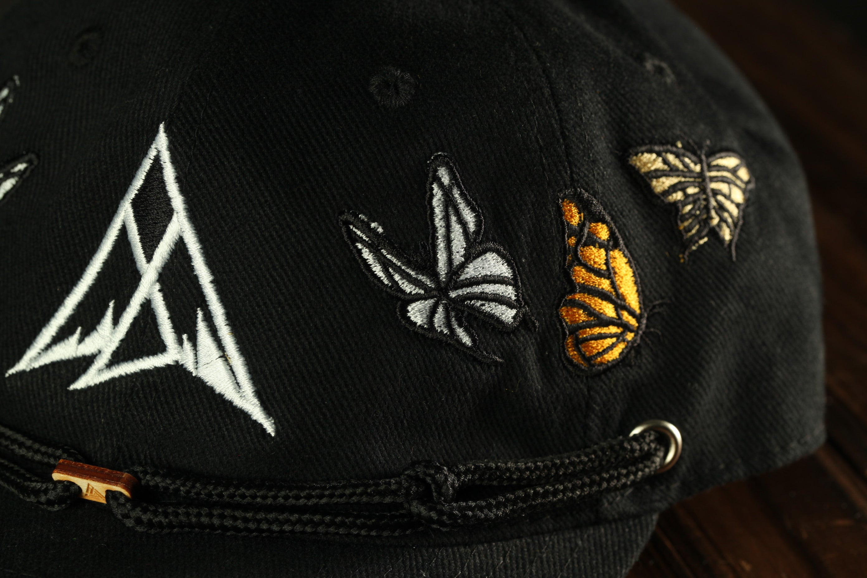 A close-up view of a black baseball cap with various embroidered designs, including a triangle, butterflies, and a butterfly with orange and black wings.