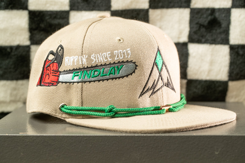 A beige baseball cap with a green string and a logo on the front, placed on a gray surface against a black and white checkered background.