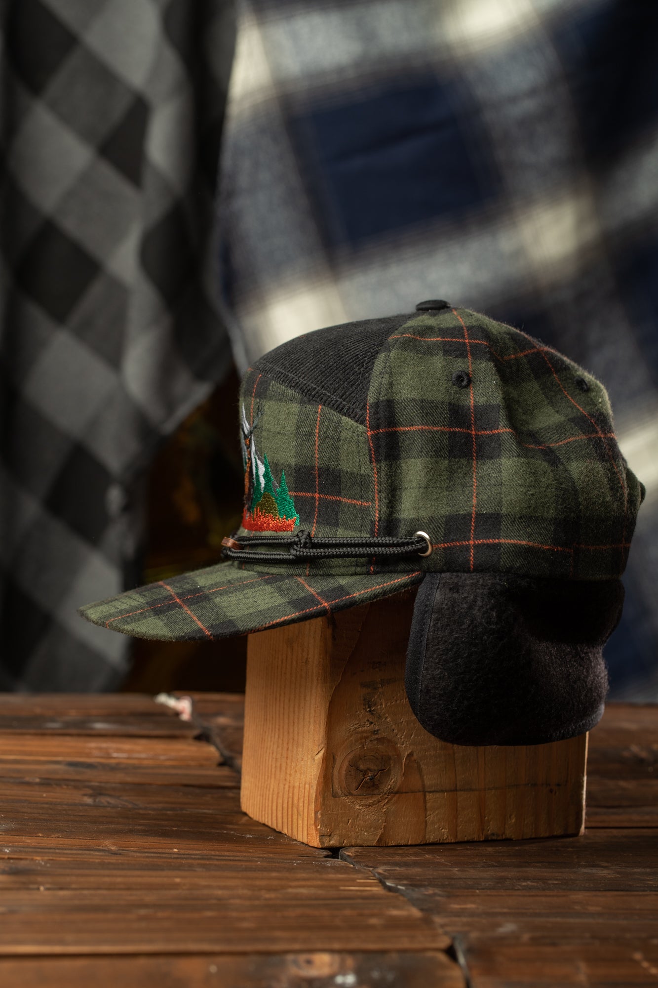 A plaid baseball cap with a black brim and a small embroidered logo on the front, displayed on a wooden stand against a blurred background.