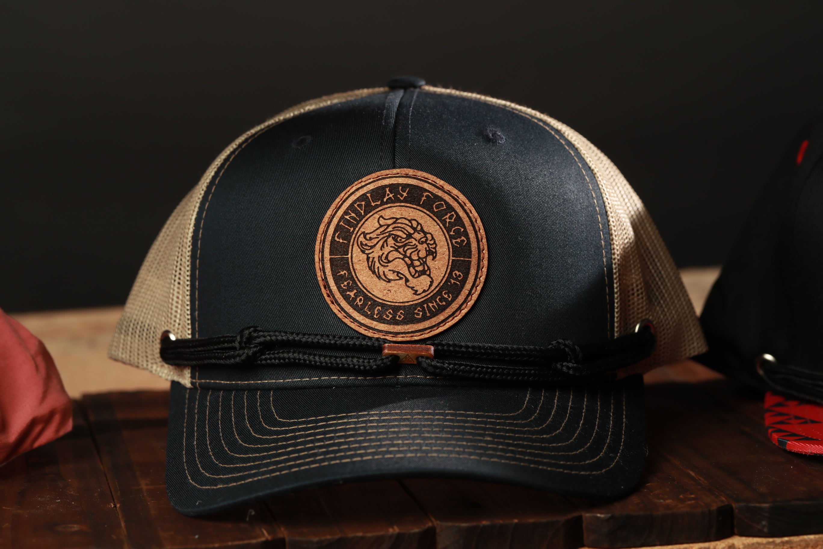A black baseball cap with a circular logo on the front, featuring a lion's head design, is displayed on a wooden surface.