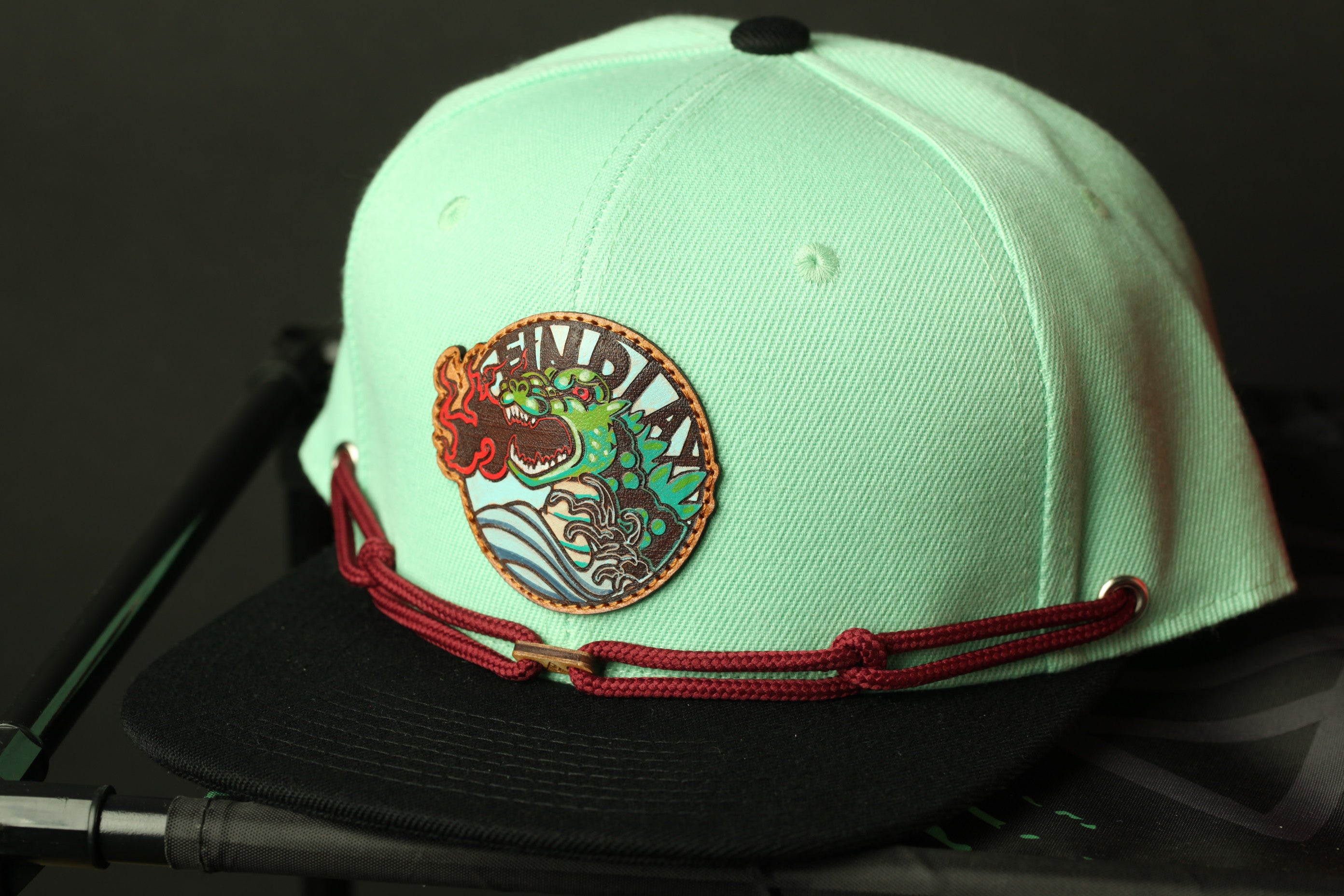 A mint green baseball cap with a red and black braided strap and a circular patch on the front.