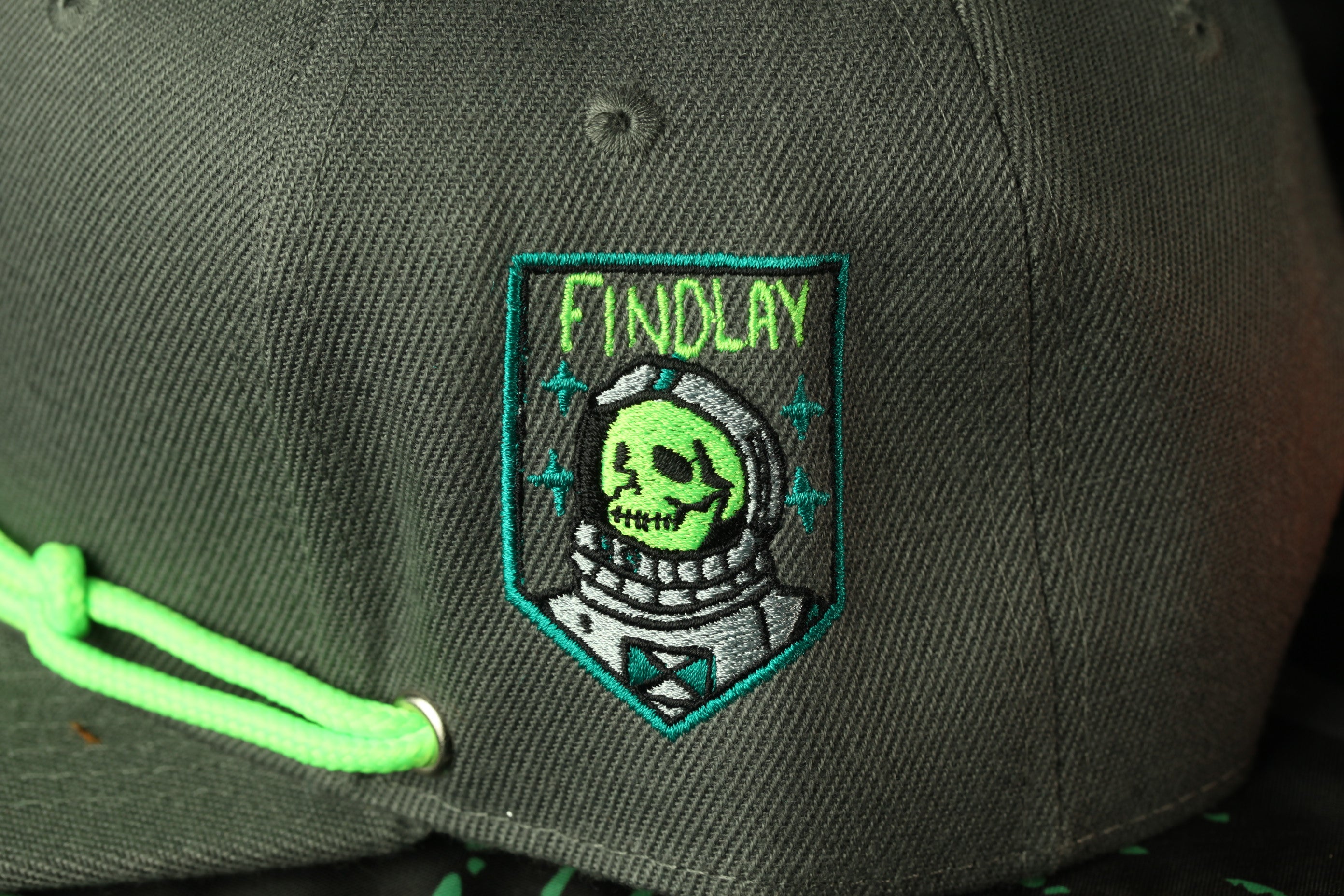 a close-up view of a dark green baseball cap with a unique embroidered patch on the front. The patch depicts a skull wearing a spacesuit, and the word "FINDLAY" is written above the skull.