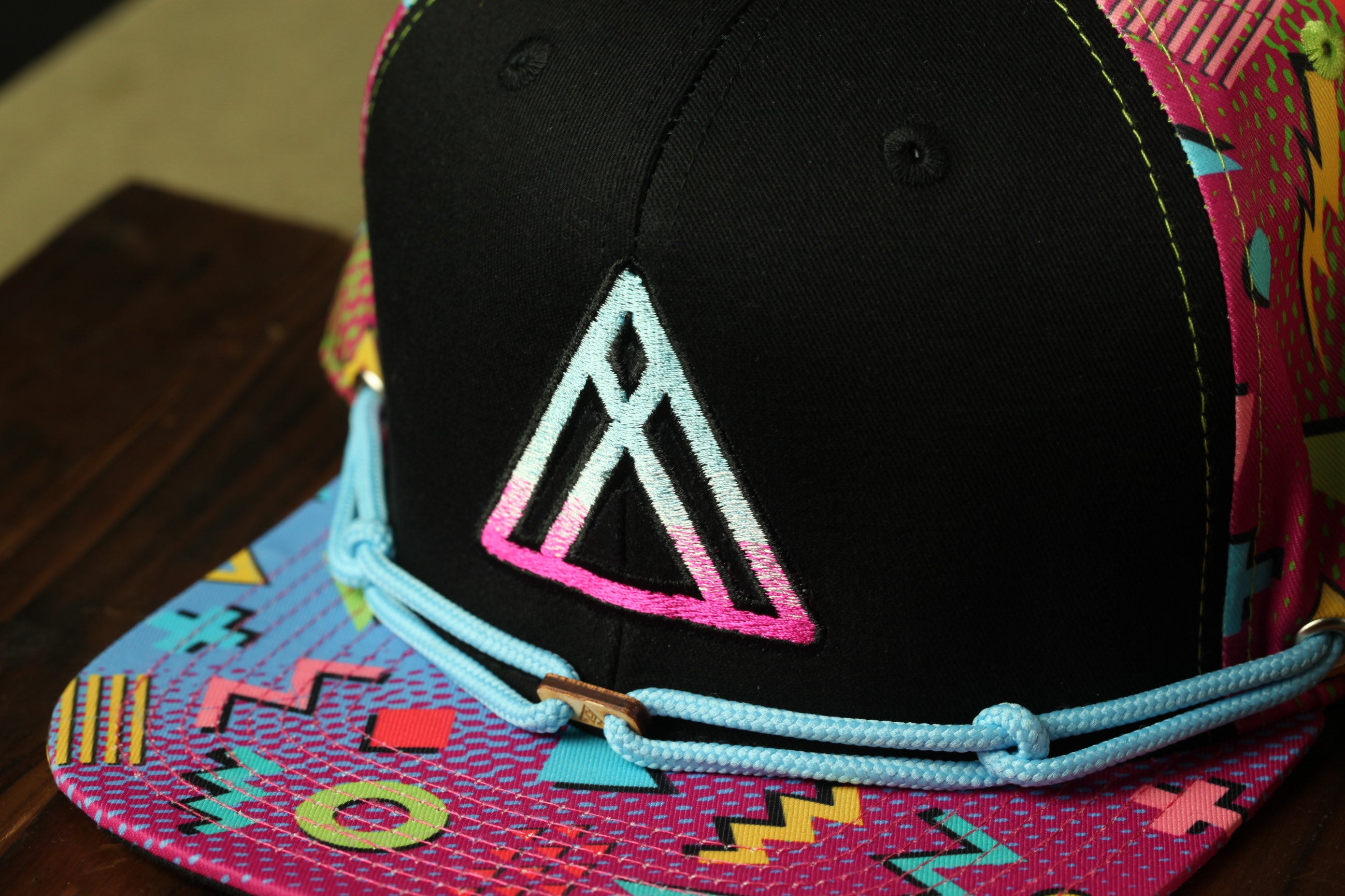 A colorful, patterned baseball cap with a black brim and a pink triangle logo on the front.