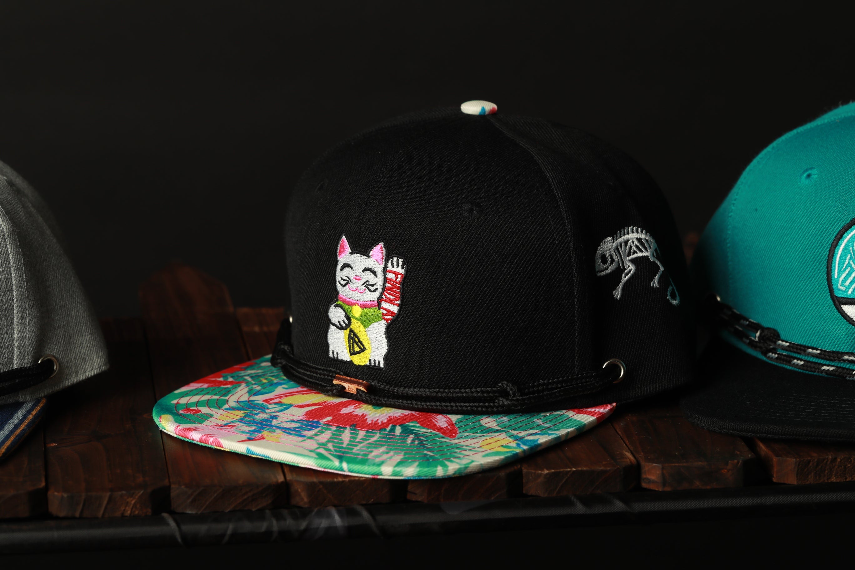 A close-up view of a black baseball cap with a colorful floral pattern and a cat embroidered on it, placed on a wooden surface.