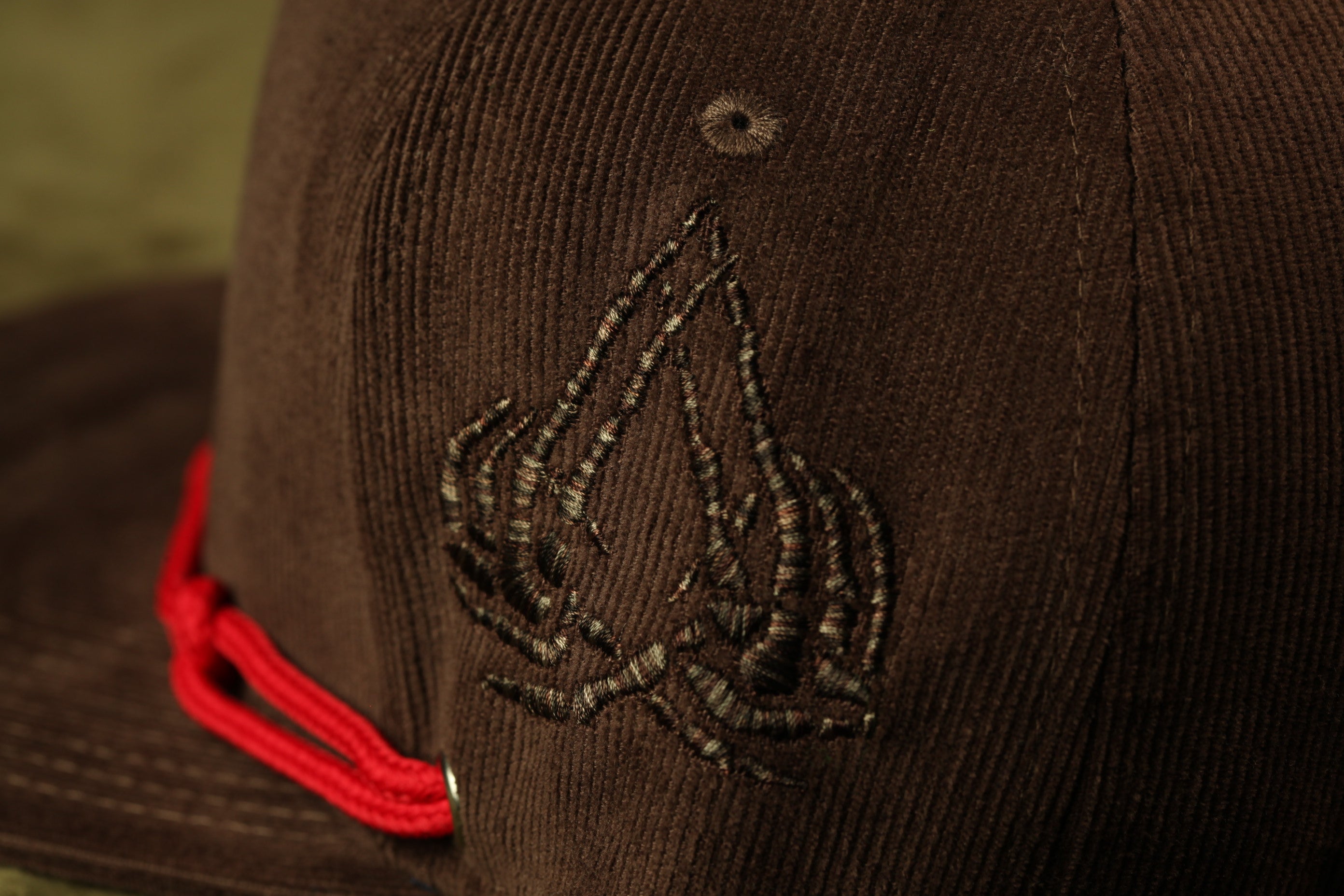 A close-up view of a brown baseball cap with a red string and a dark, intricate embroidered design on the front.