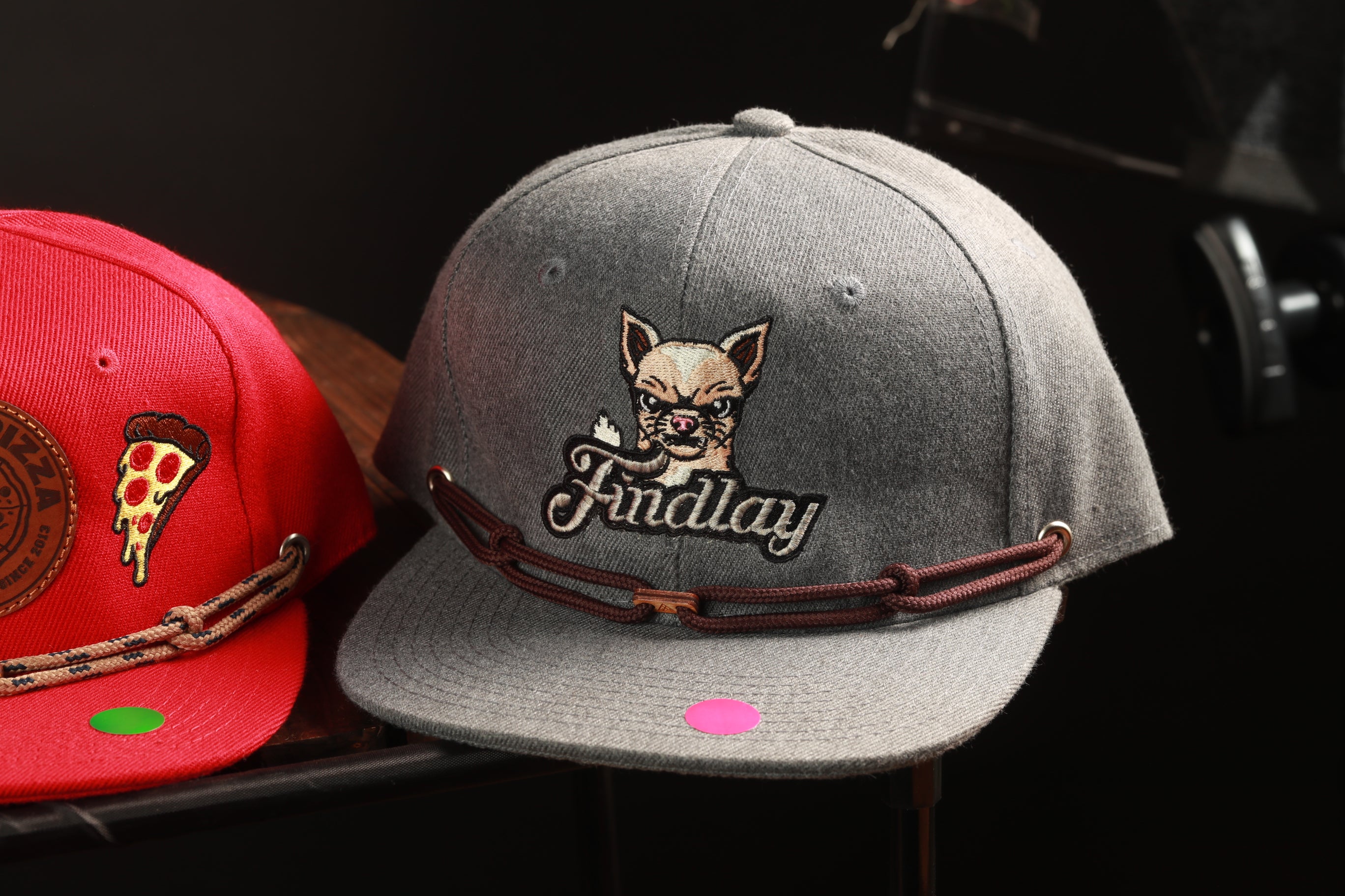 two baseball caps, one gray and one red, with the gray cap displaying a cartoon dog and the red cap having a pizza slice design.