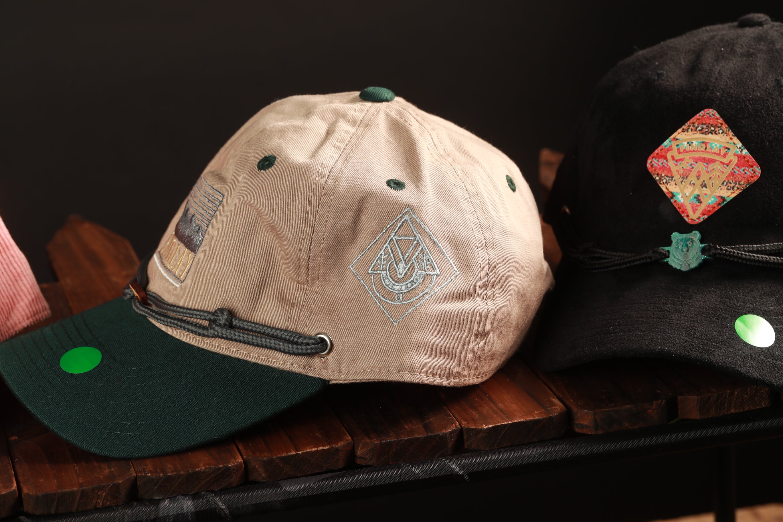 Two baseball caps are displayed on a wooden surface, one beige and one black, with green and red designs on them.