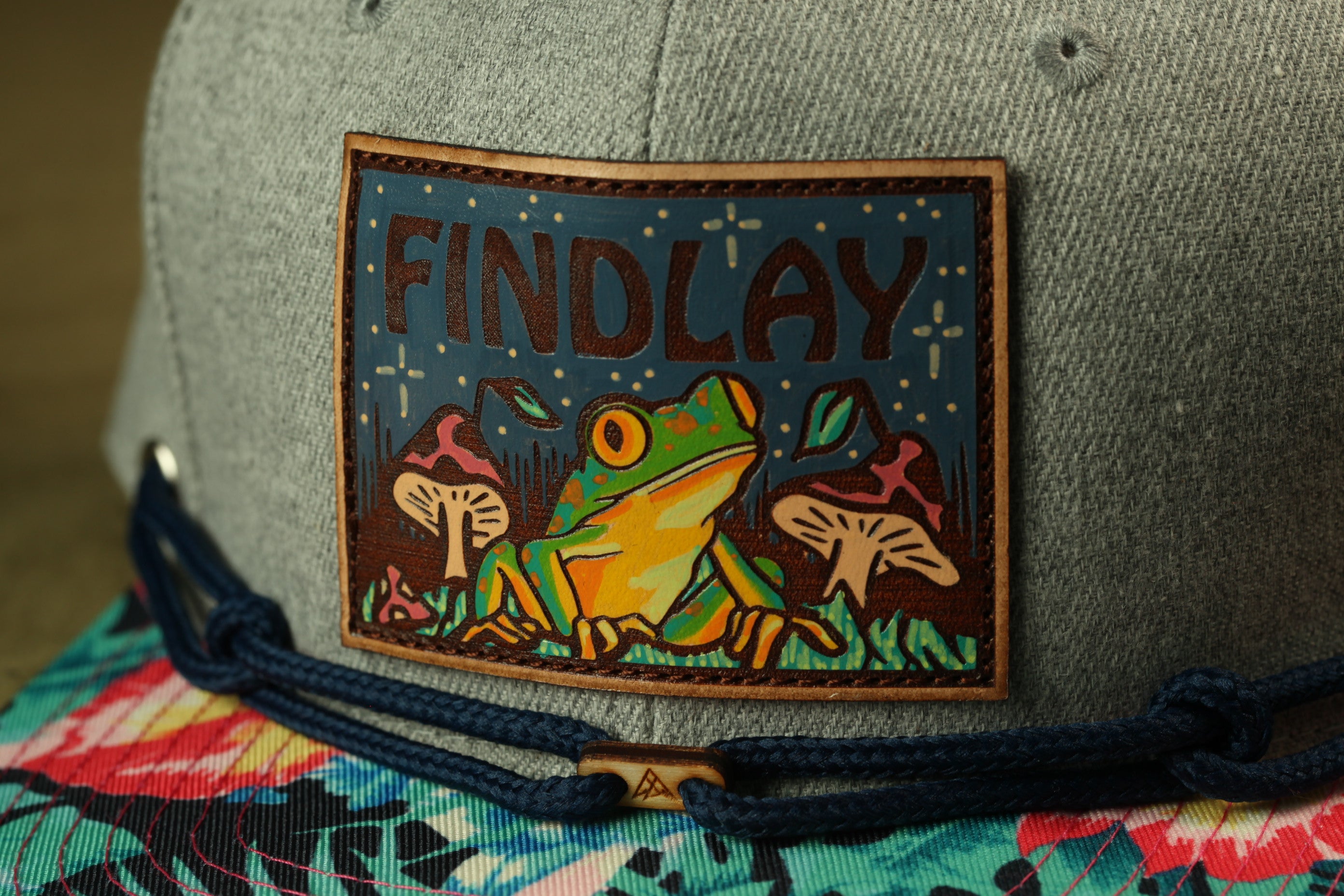 A gray baseball cap with a patch featuring a frog and mushrooms, along with the text "FINDLAY".