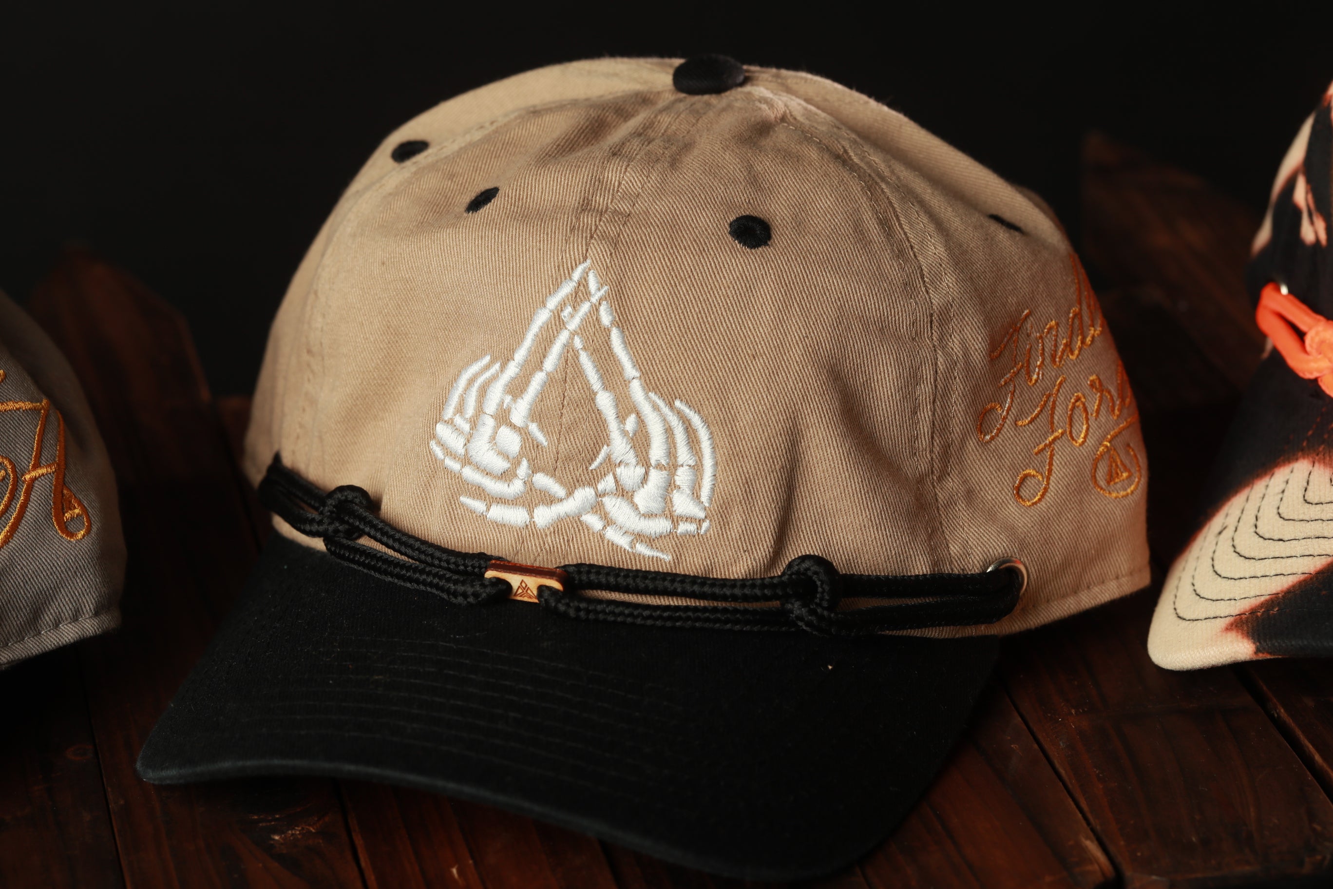 A beige baseball cap with a black brim and a white graphic design on the front, sitting on a wooden surface.