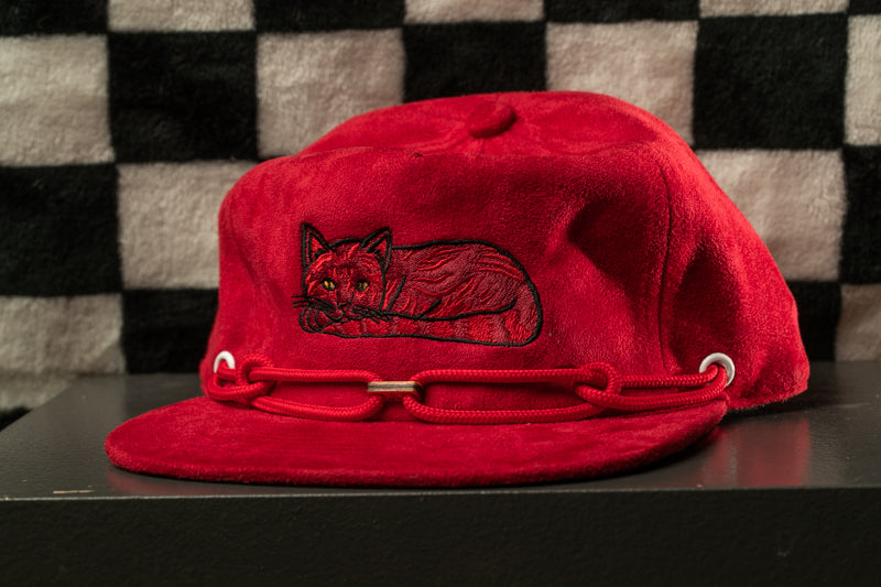 A red baseball cap with a cat embroidered on it, sitting on a black surface against a checkered background.