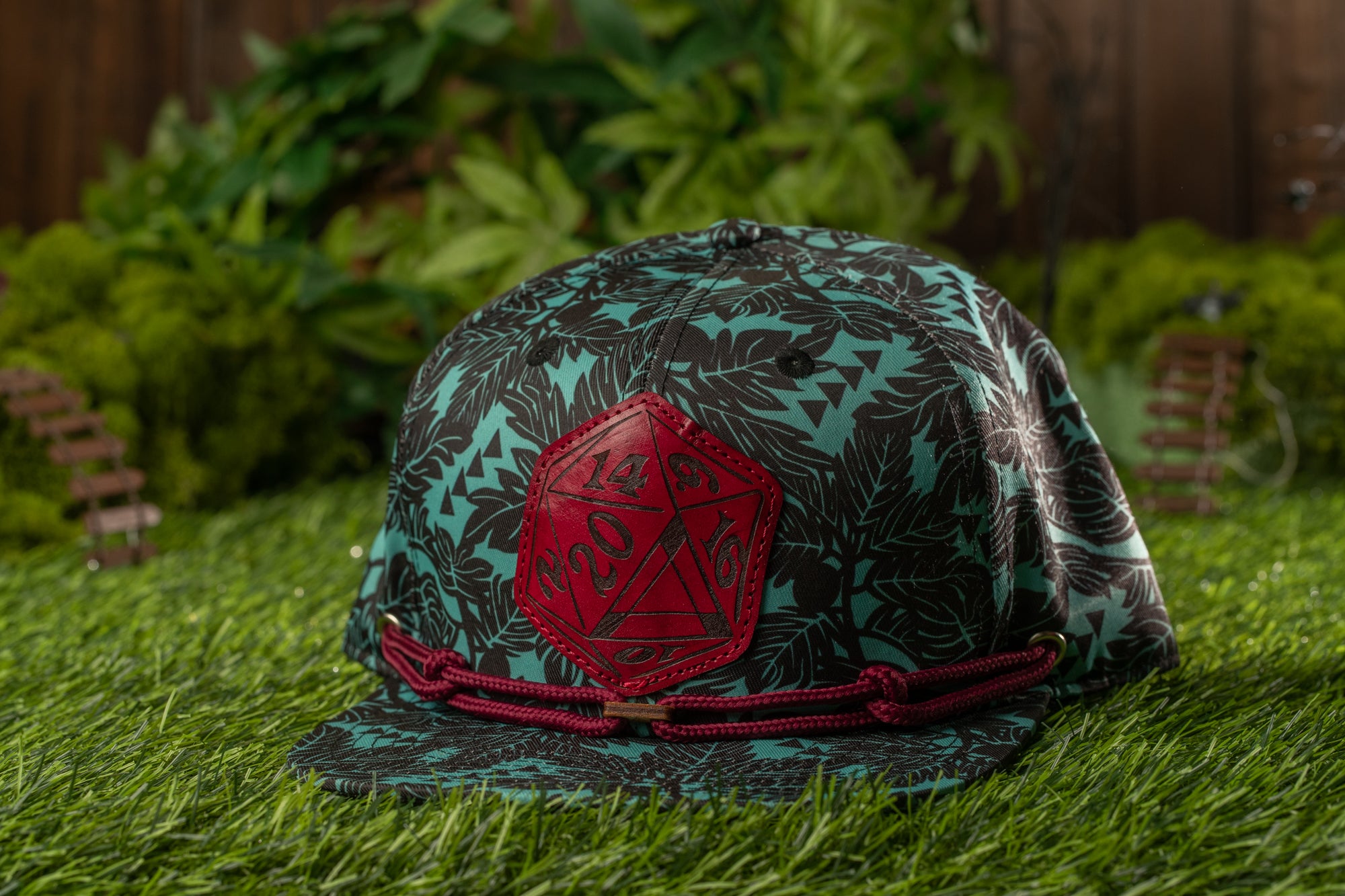 A green baseball cap with a red hexagon patch on the front, placed on a grassy surface with a blurred background of greenery.