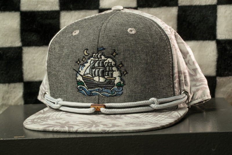A gray baseball cap with a printed design of a ship on the front, sitting on a black surface against a checkered background.