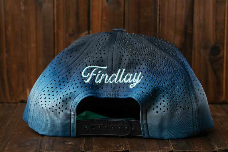 A blue baseball cap with the word "Findlay" embroidered on it, sitting on a wooden surface.