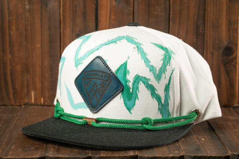 A white baseball cap with a green and black design, featuring a logo on the front, is displayed on a wooden surface.