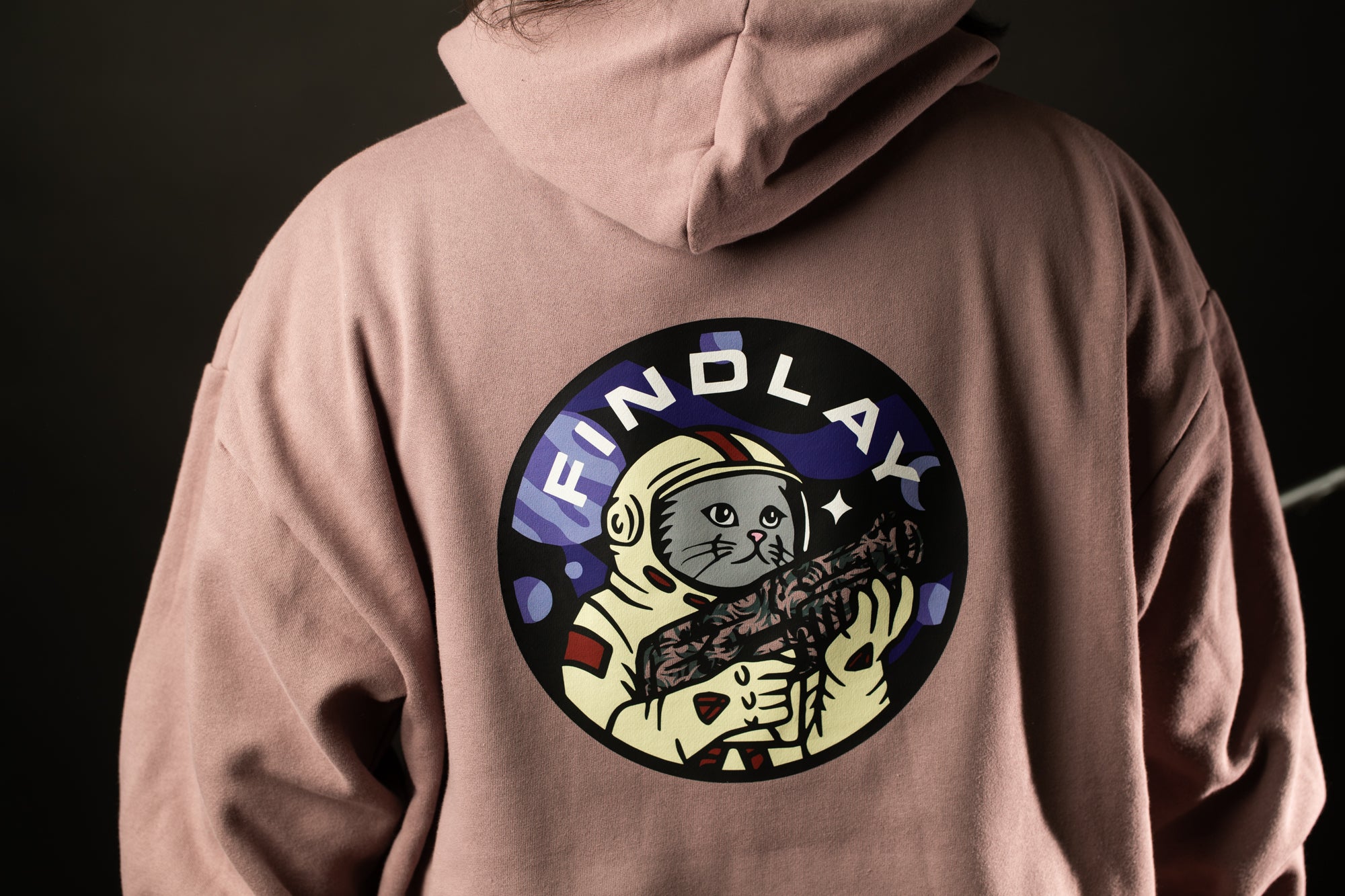 A person wearing a pink hoodie with a circular graphic design featuring a cat wearing a spacesuit and holding a rocket ship.