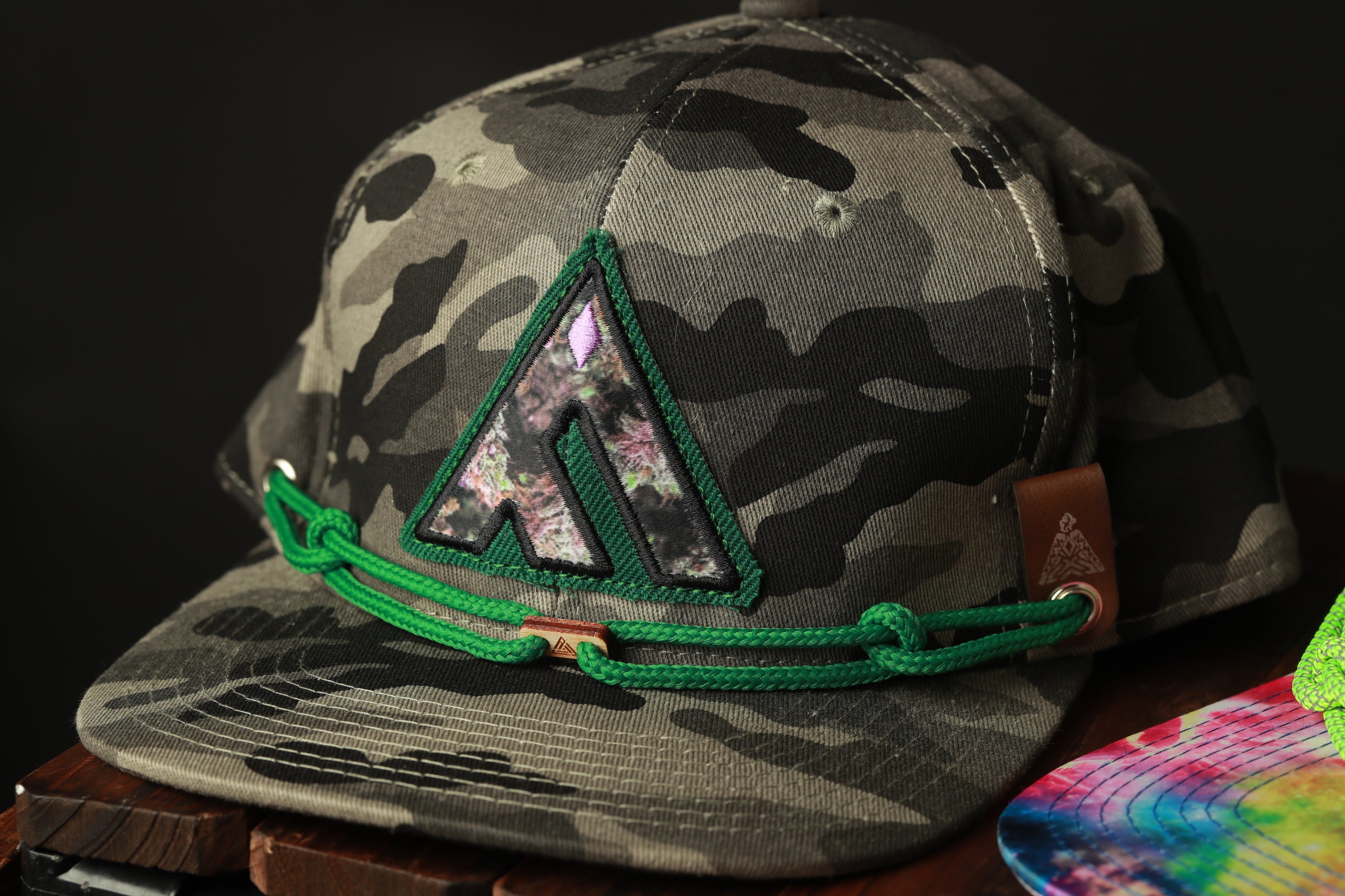 A camouflage baseball cap with a green chain and a small logo on the front, placed on a wooden surface.