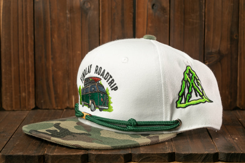 A white baseball cap with a camouflage pattern and a graphic of a van, along with the text "FIND A ROAD TRIP" printed on it, is displayed on a wooden surface.
