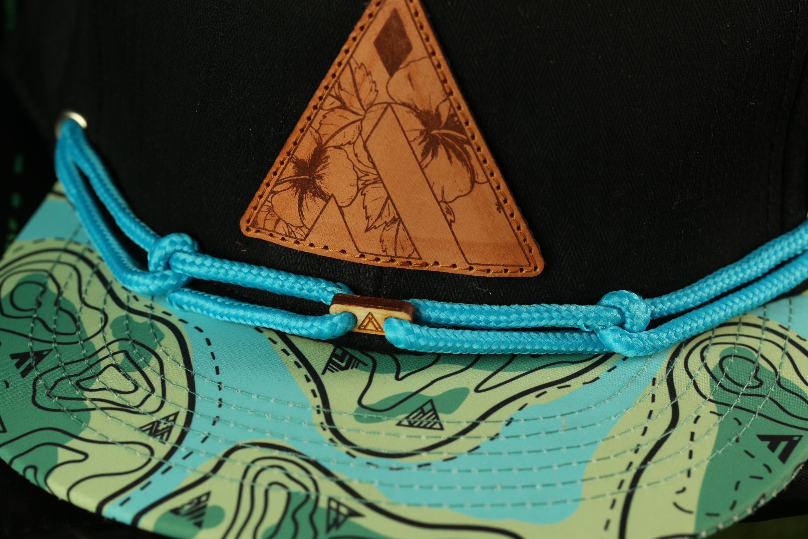 a close-up view of a black baseball cap with a distinctive triangular patch featuring a floral design. The cap has a blue string and a black strap, and the cap is set against a background with a map pattern.